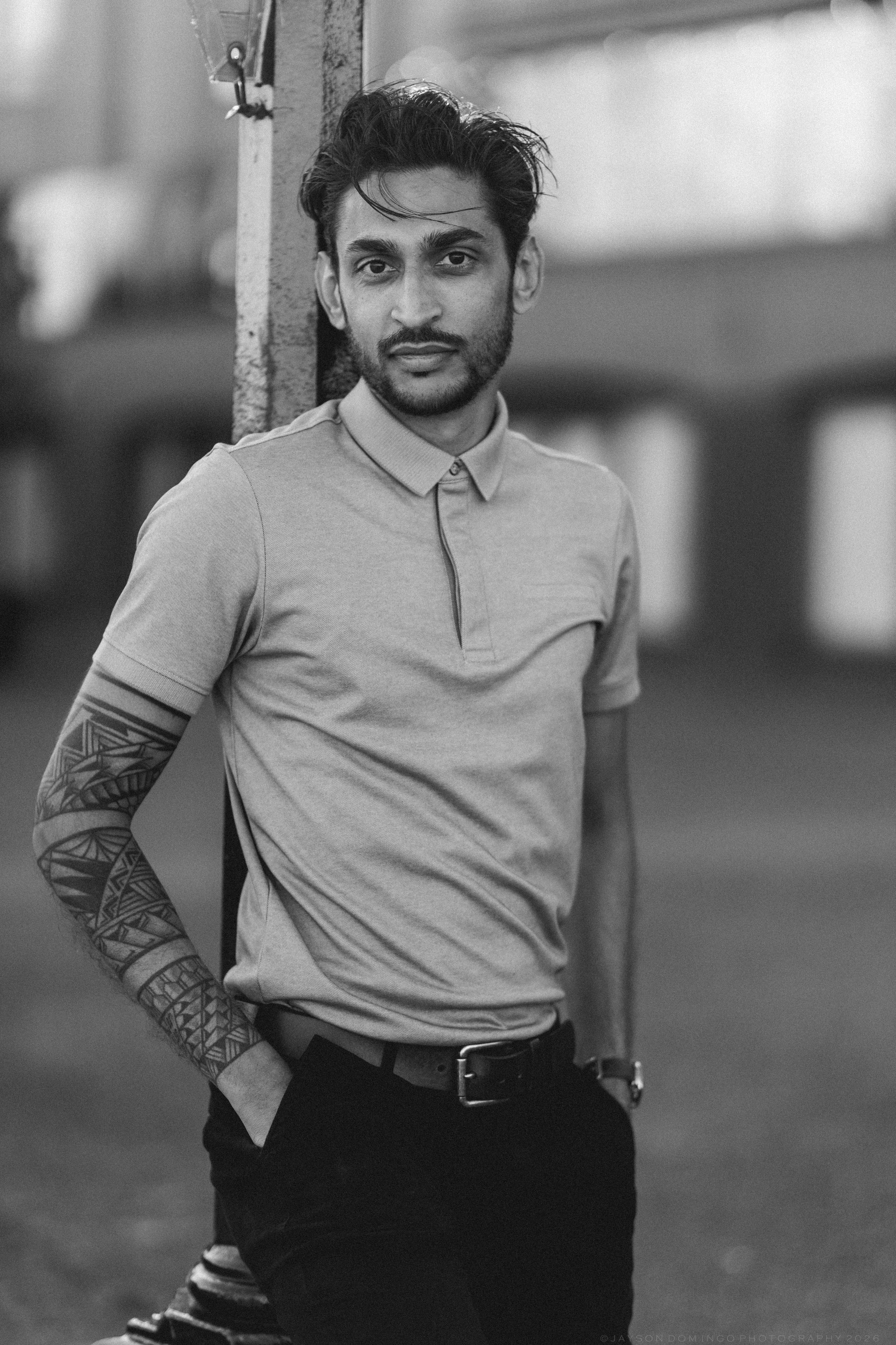 Black and white close-up portrait of a tattooed man leaning against a wall outdoors.