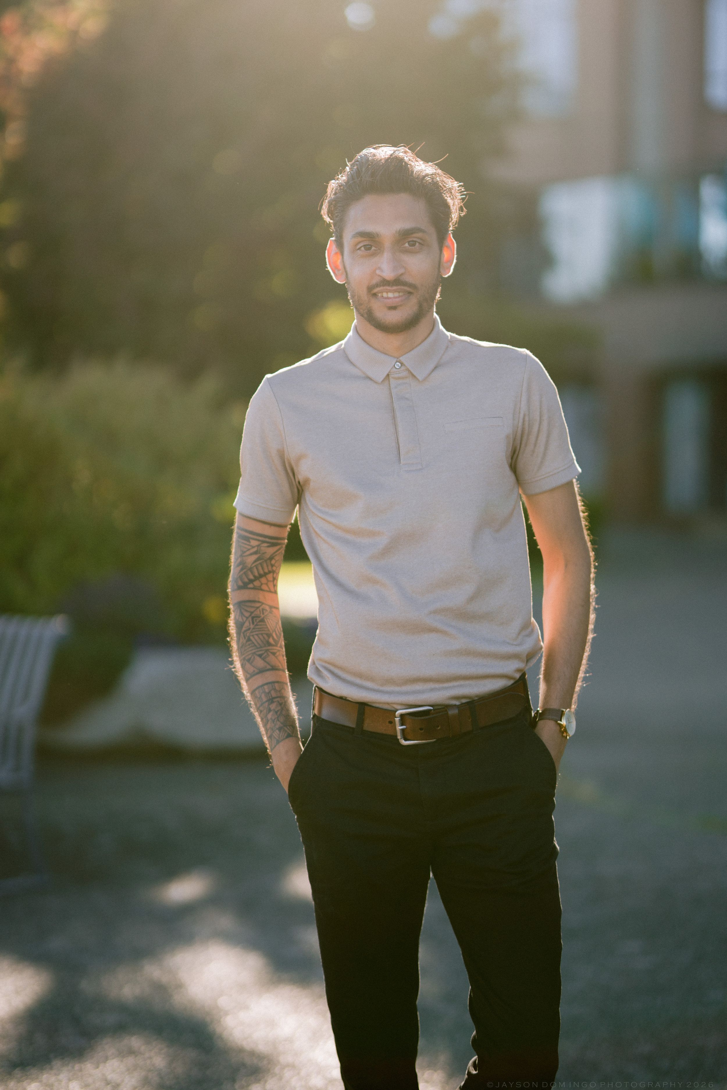 Smiling tattooed man in a polo shirt standing in a sunlit outdoor garden setting.