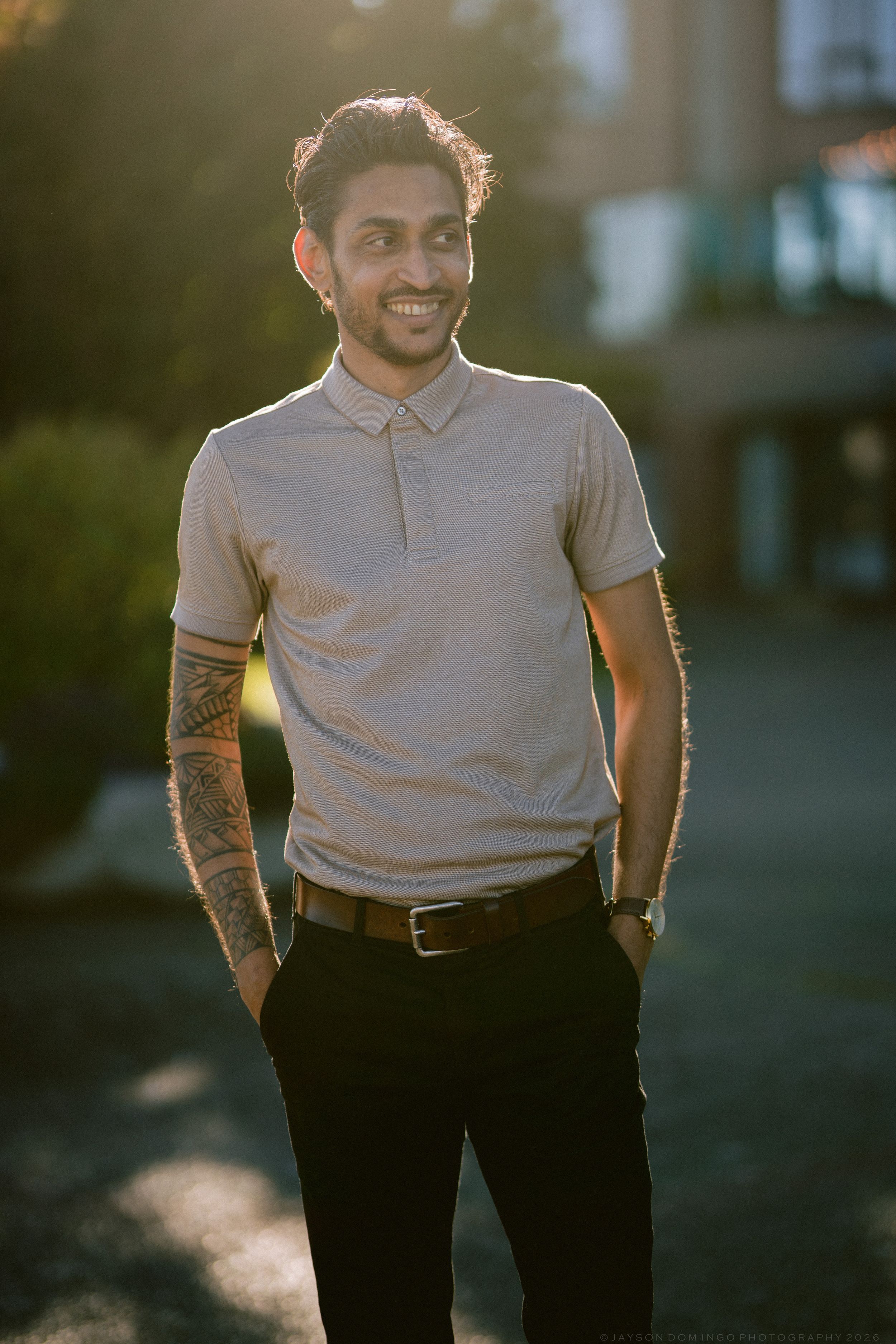 Smiling man with tattoos wearing grey polo shirt standing outdoors in warm golden hour sunlight.