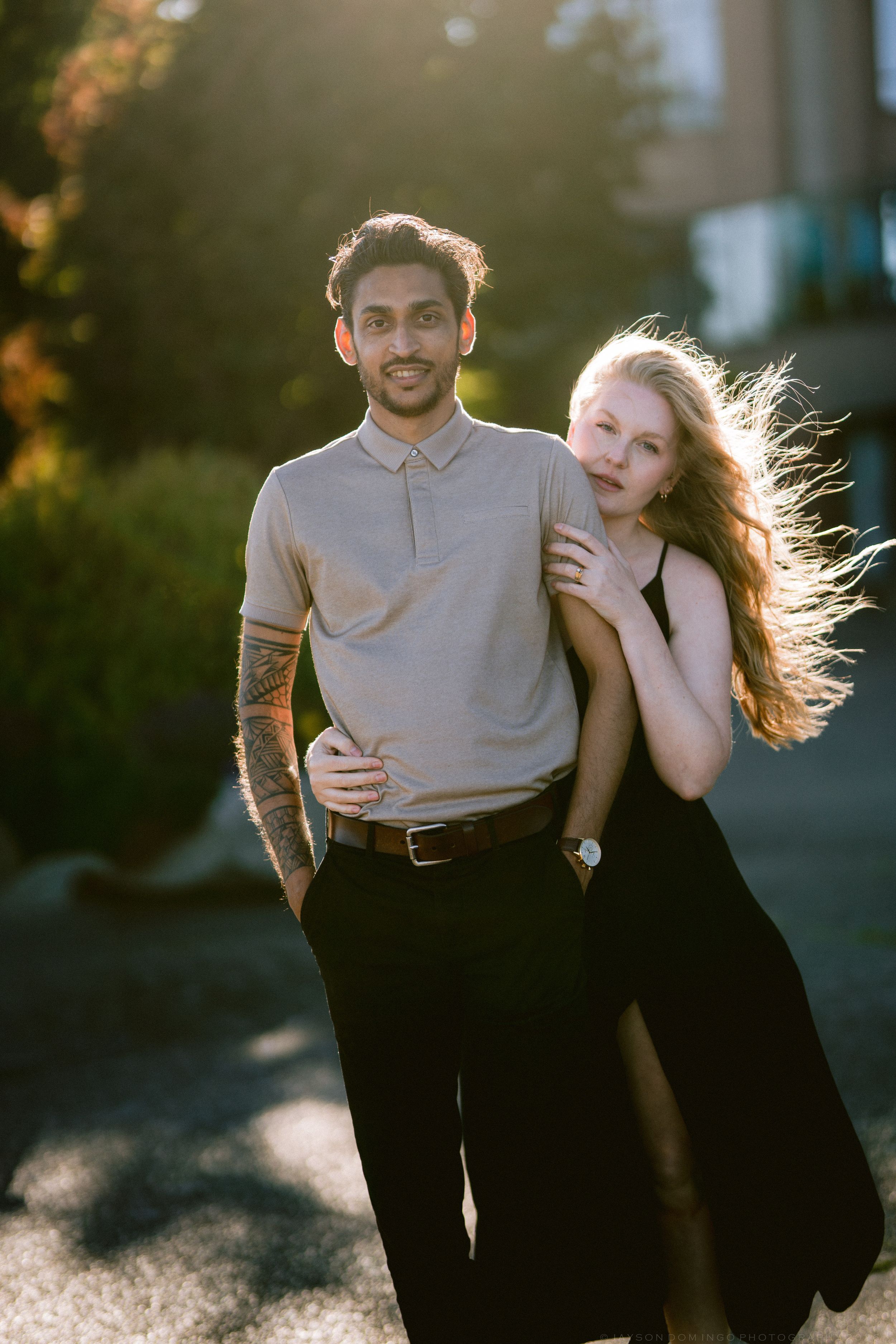 Couple posing outdoors in golden hour light, man with tattoos in grey polo embracing blonde woman in black dress.
