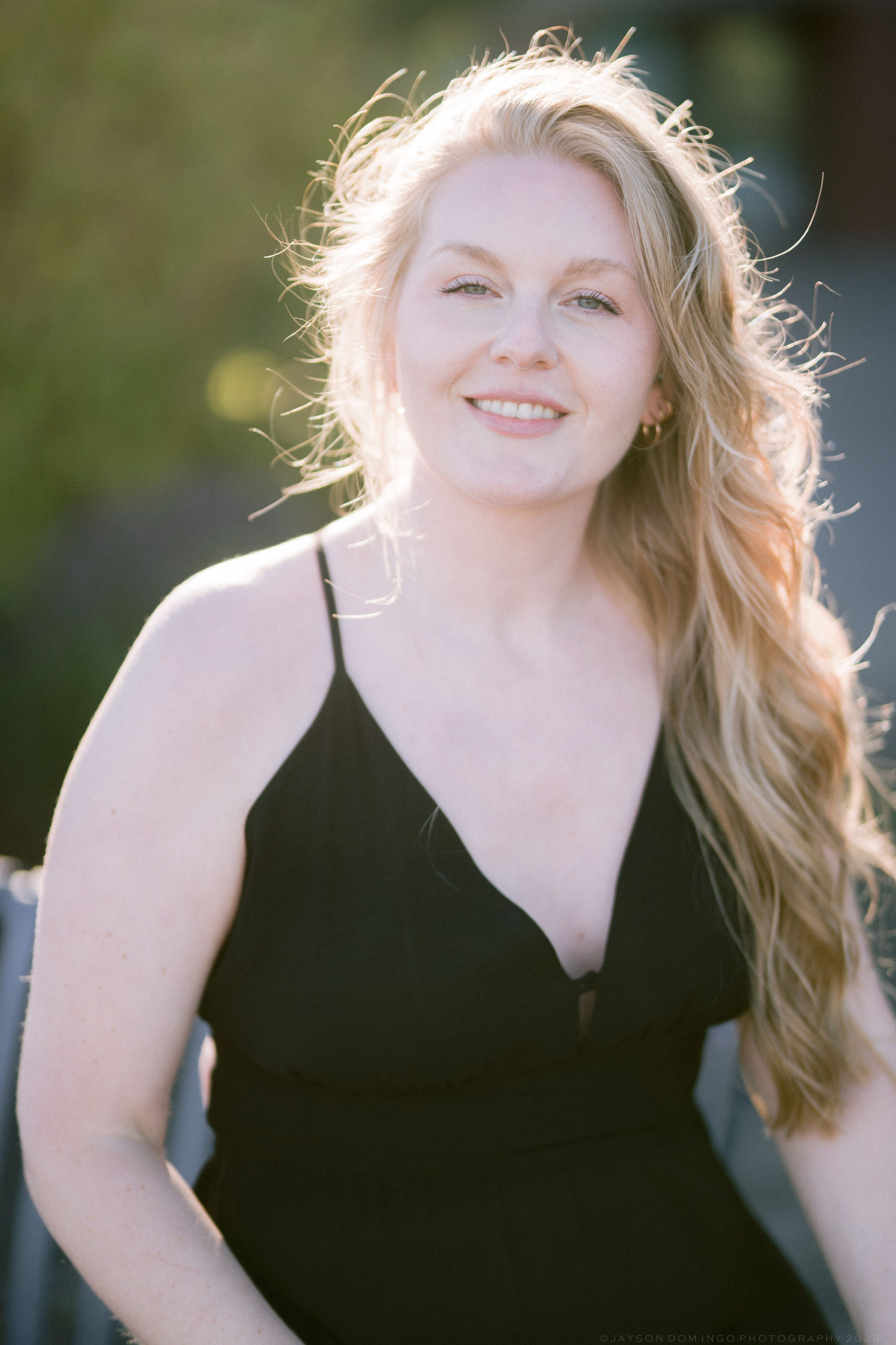 Smiling blonde woman in black dress posing in warm golden sunlight.