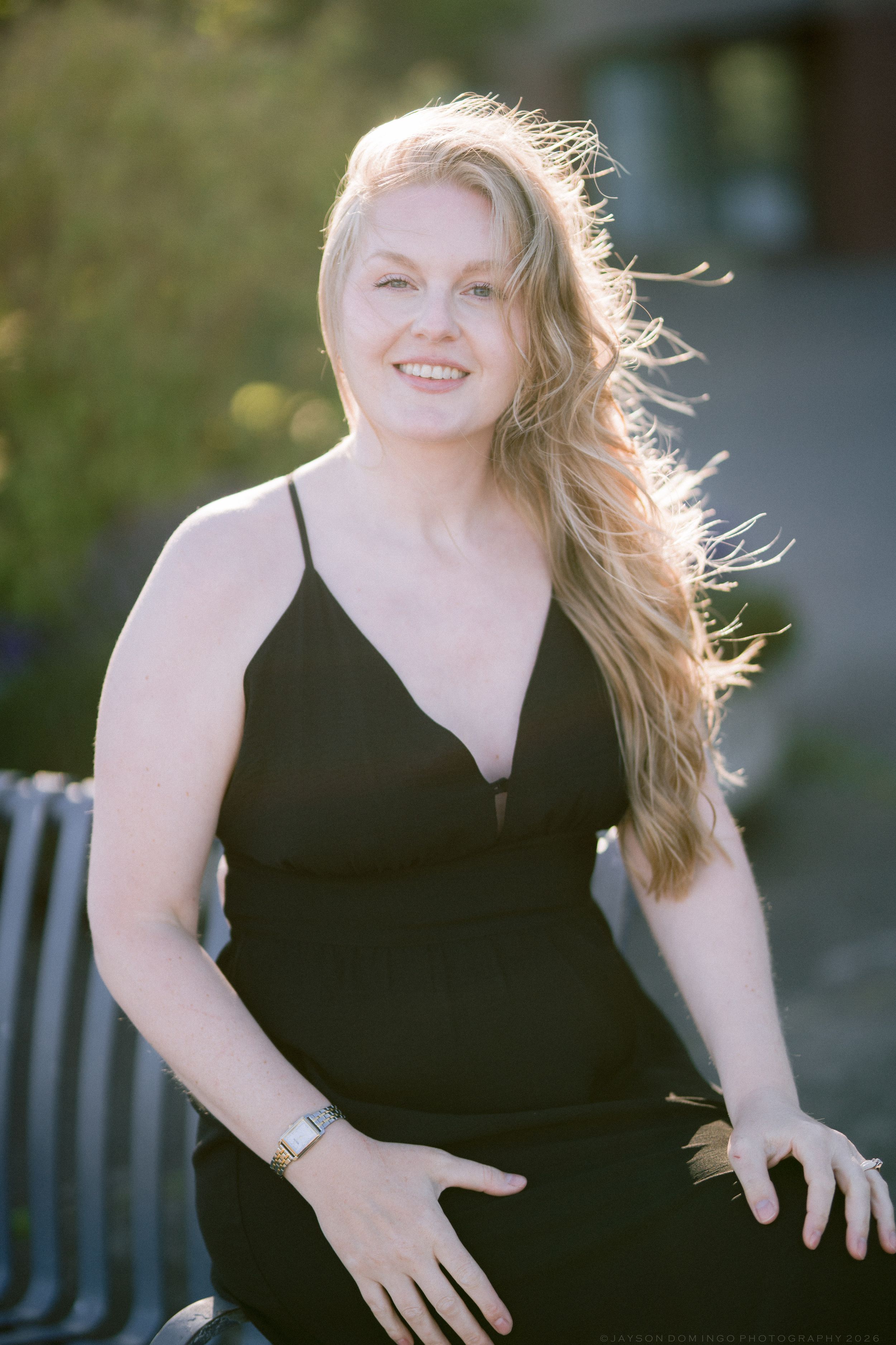 Smiling blonde woman in black dress sitting outdoors in golden sunlight with hair blowing in wind.