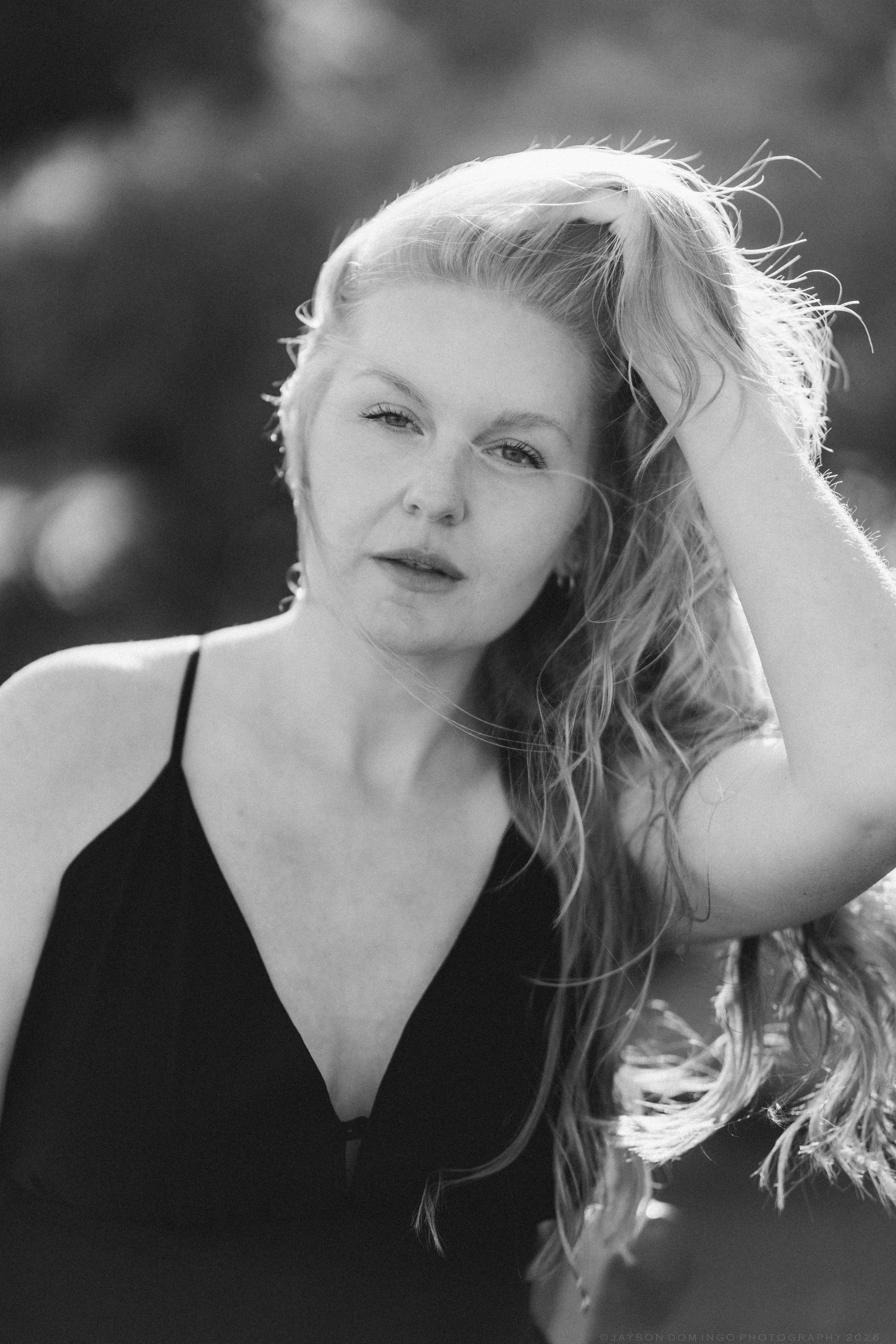 Black and white portrait of blonde woman holding her hair back, wearing black dress in natural light.