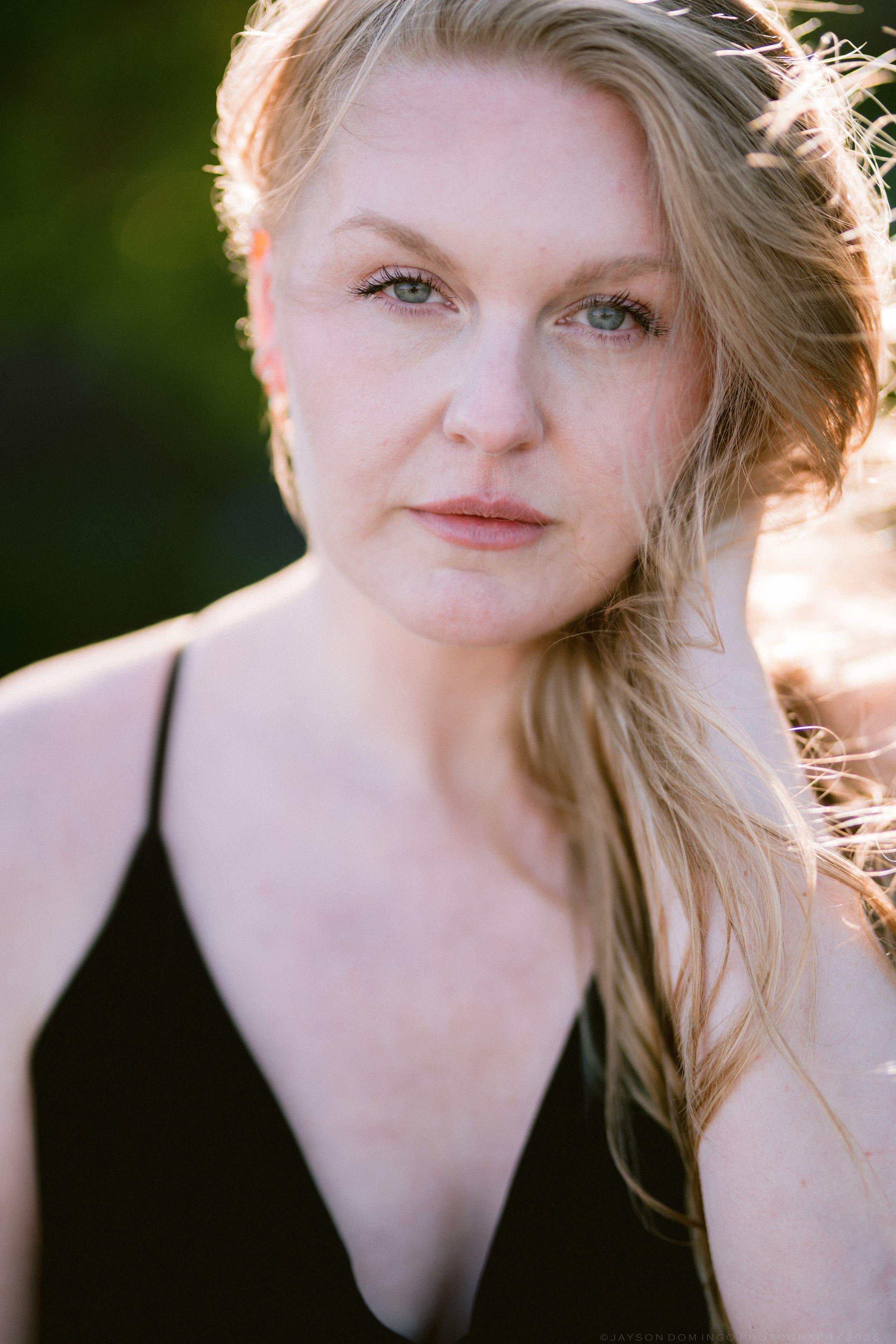 Blonde woman in black dress with blue eyes gazing at camera in golden sunlight.