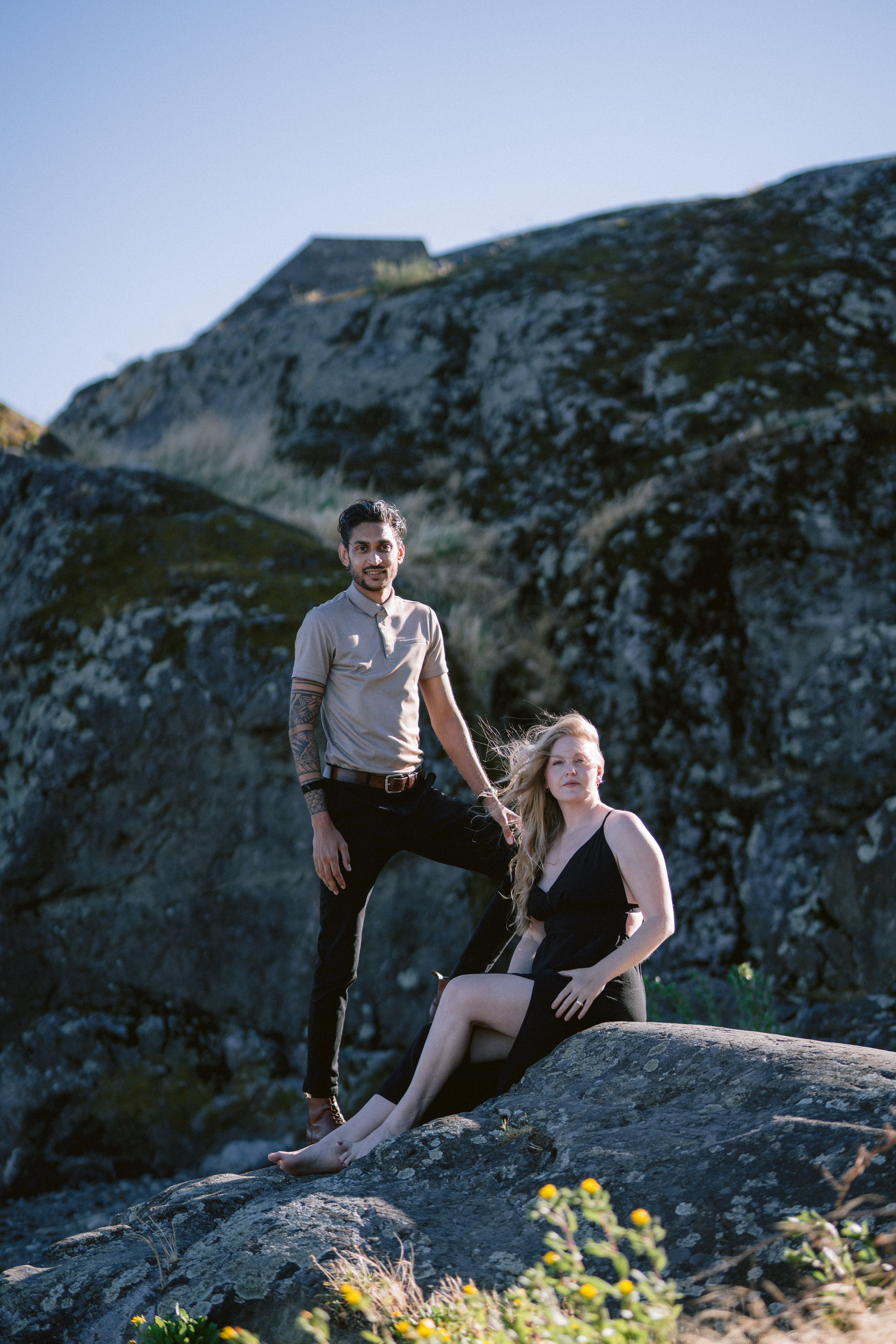 Couple posing on rocky terrain outdoors, man standing while woman sits gracefully on boulder below.