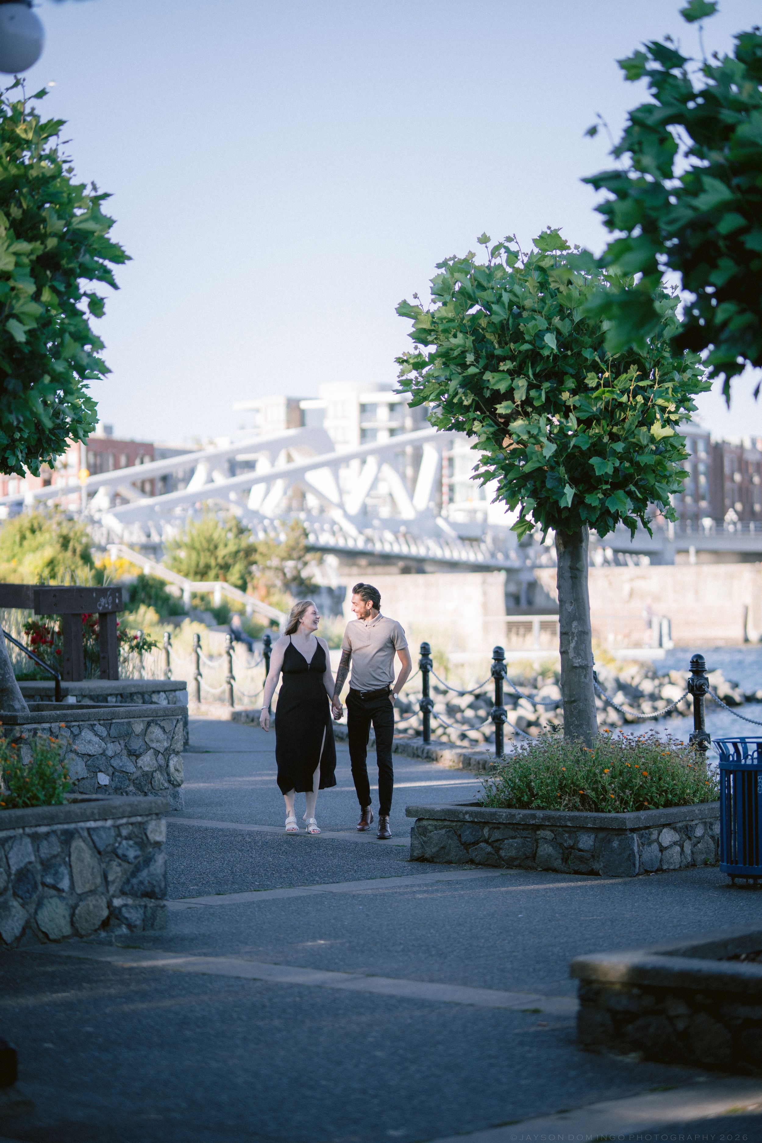 Couple walking hand in hand through a waterfront park with lush trees and bridge backdrop.