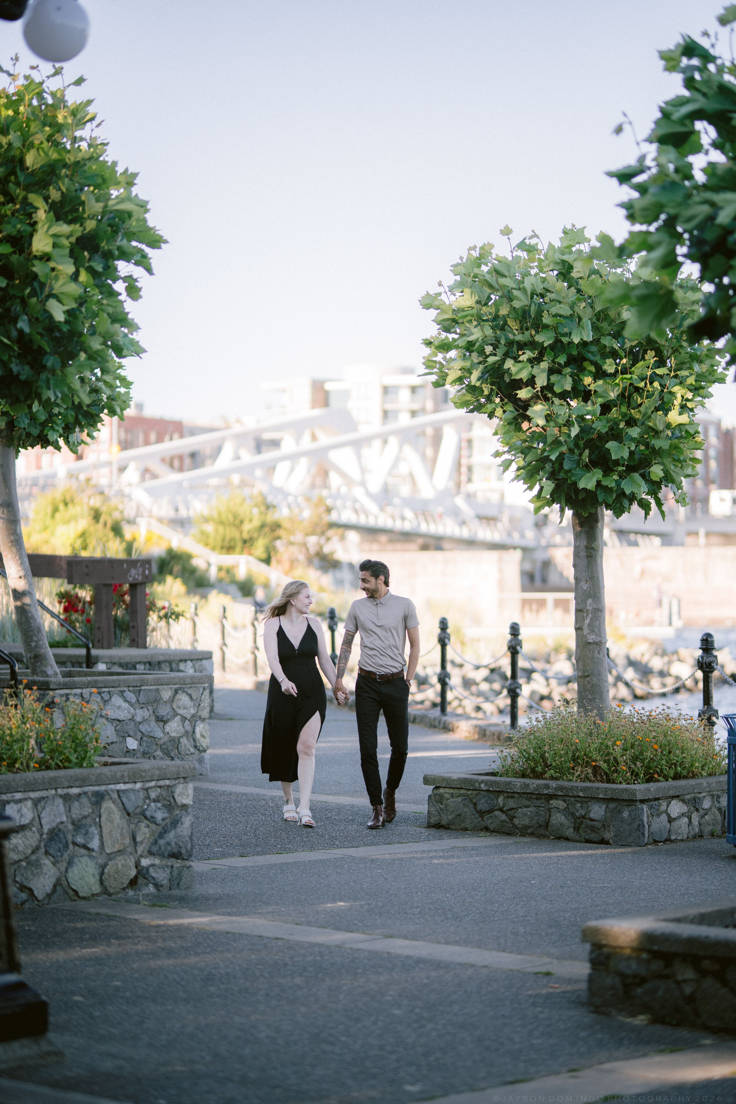 Couple holding hands walking through scenic waterfront park with bridge and trees in background.