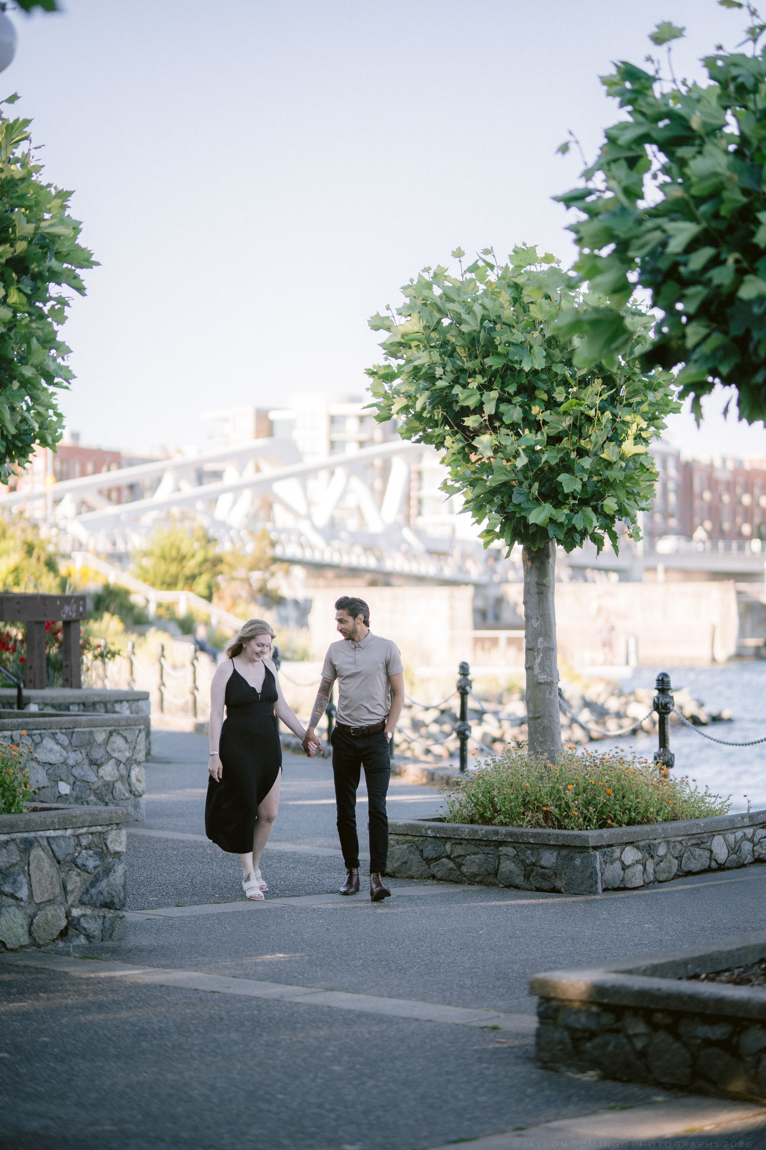 Couple strolling hand in hand along a scenic waterfront pathway with manicured trees.