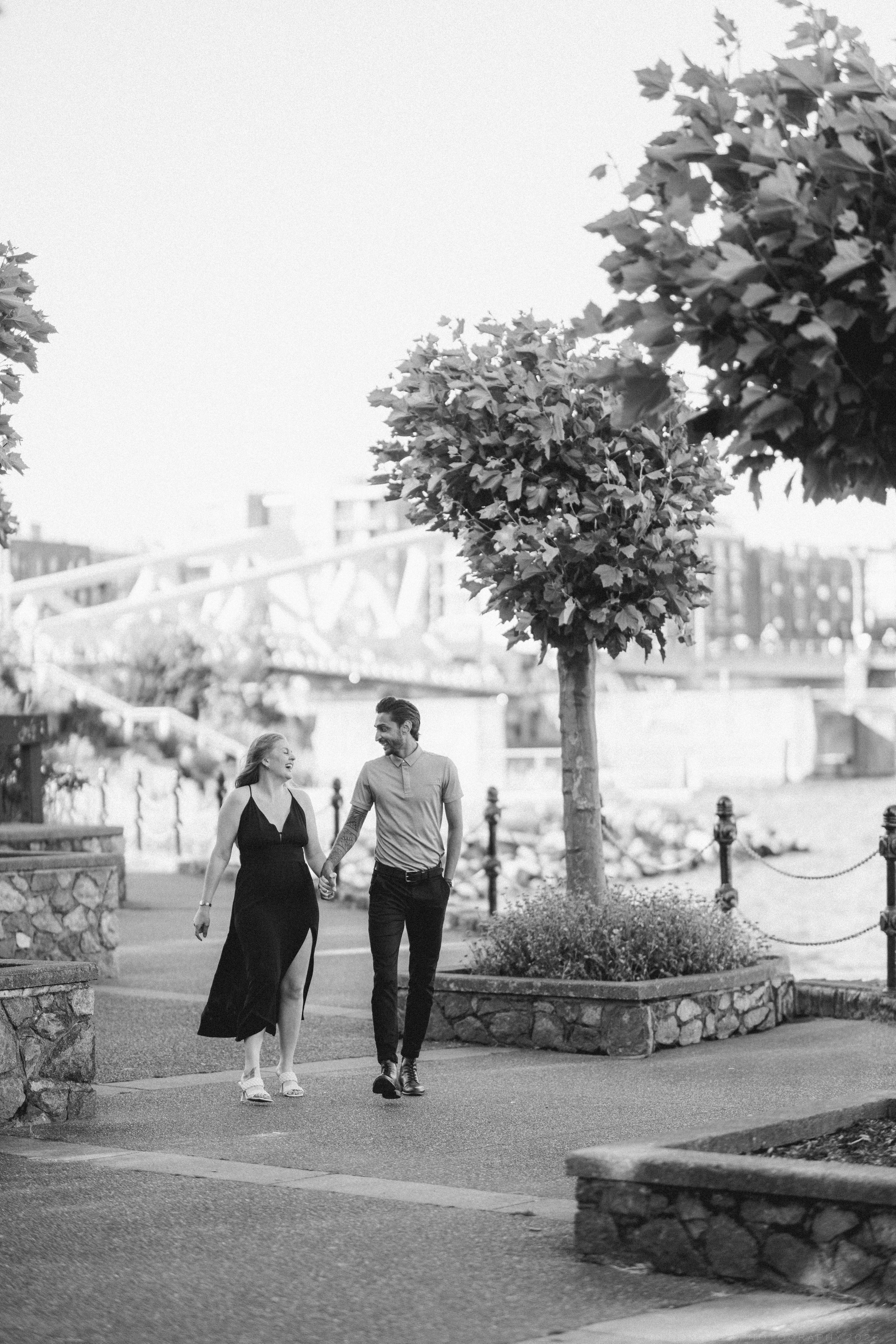 Black and white photo of a couple holding hands walking along a waterfront promenade with trees and a bridge behind.