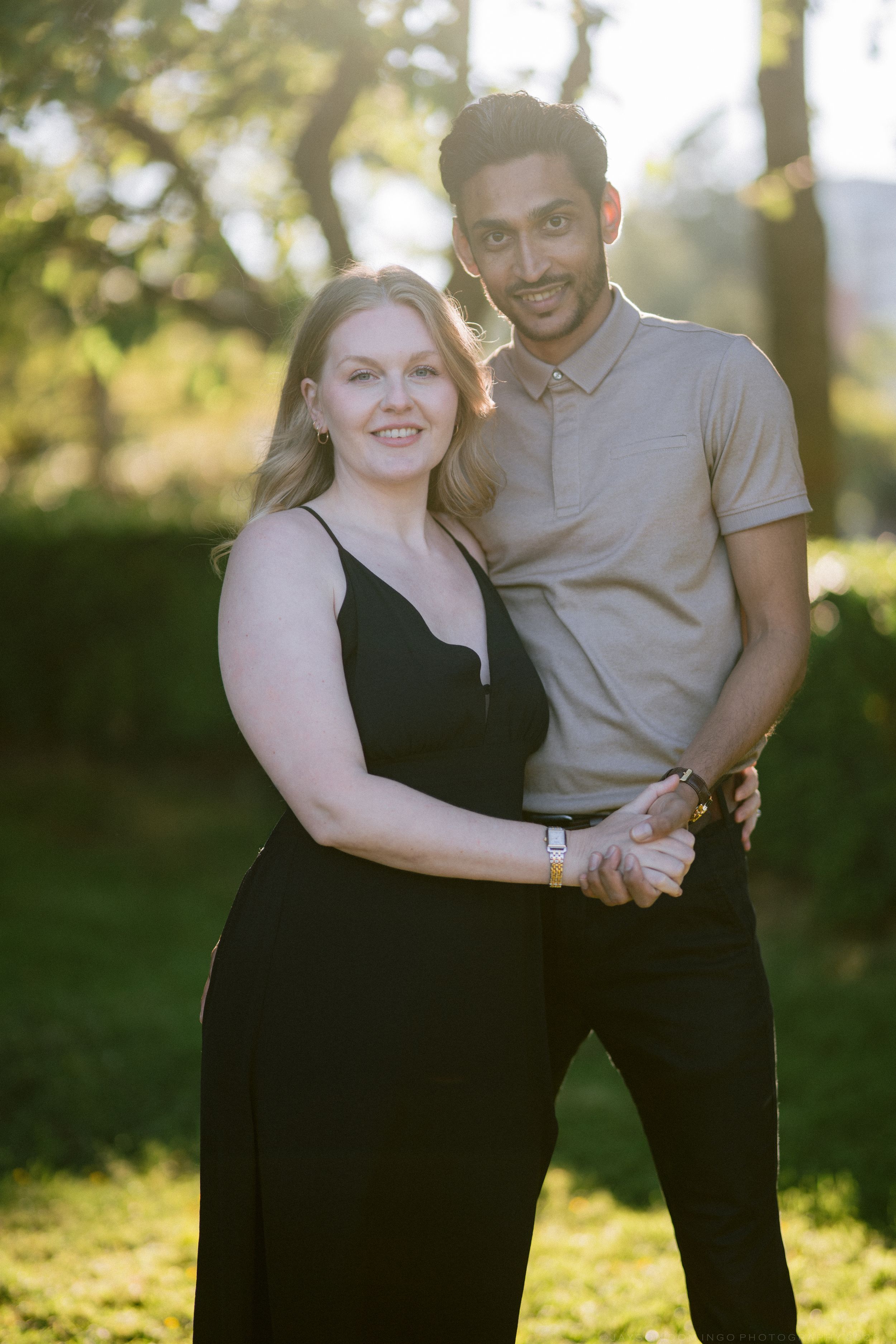 A smiling couple poses outdoors in golden sunlight, woman in black dress holding hands with man in gray polo shirt.