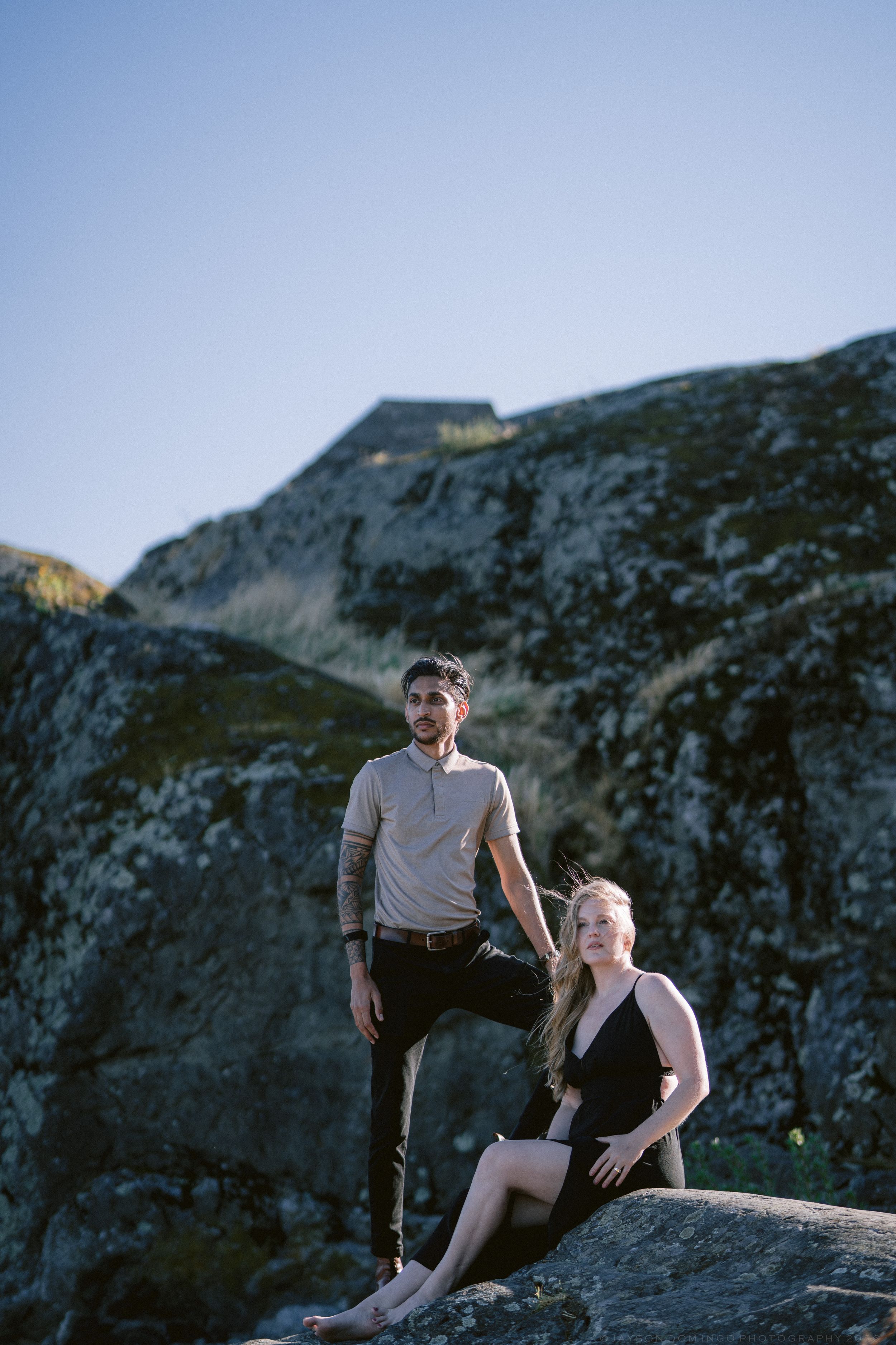 A couple poses on large rocks outdoors, man standing with foot raised while woman sits barefoot in black dress.