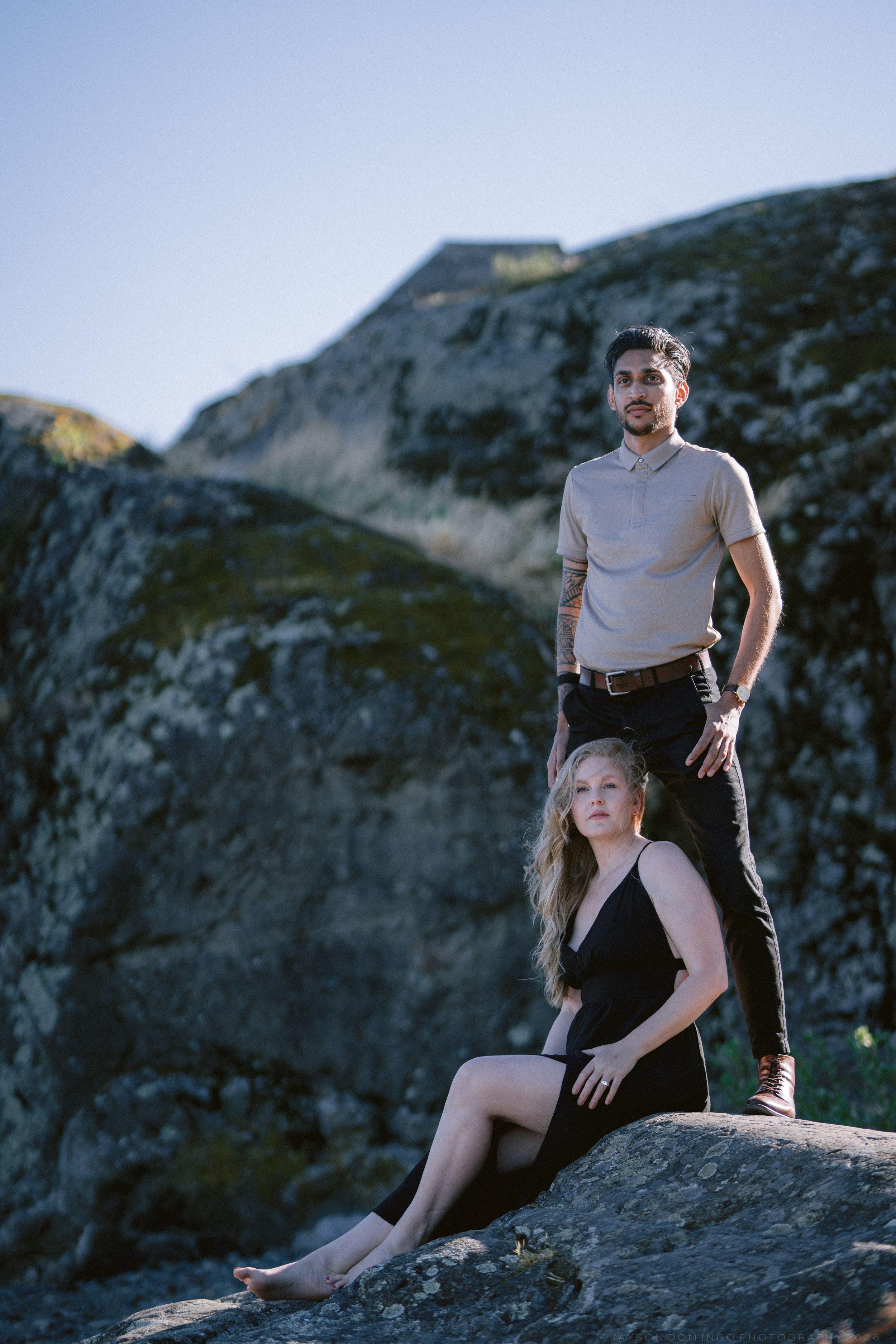 A couple poses dramatically on rocky terrain, man standing behind seated blonde woman in black dress at golden hour.