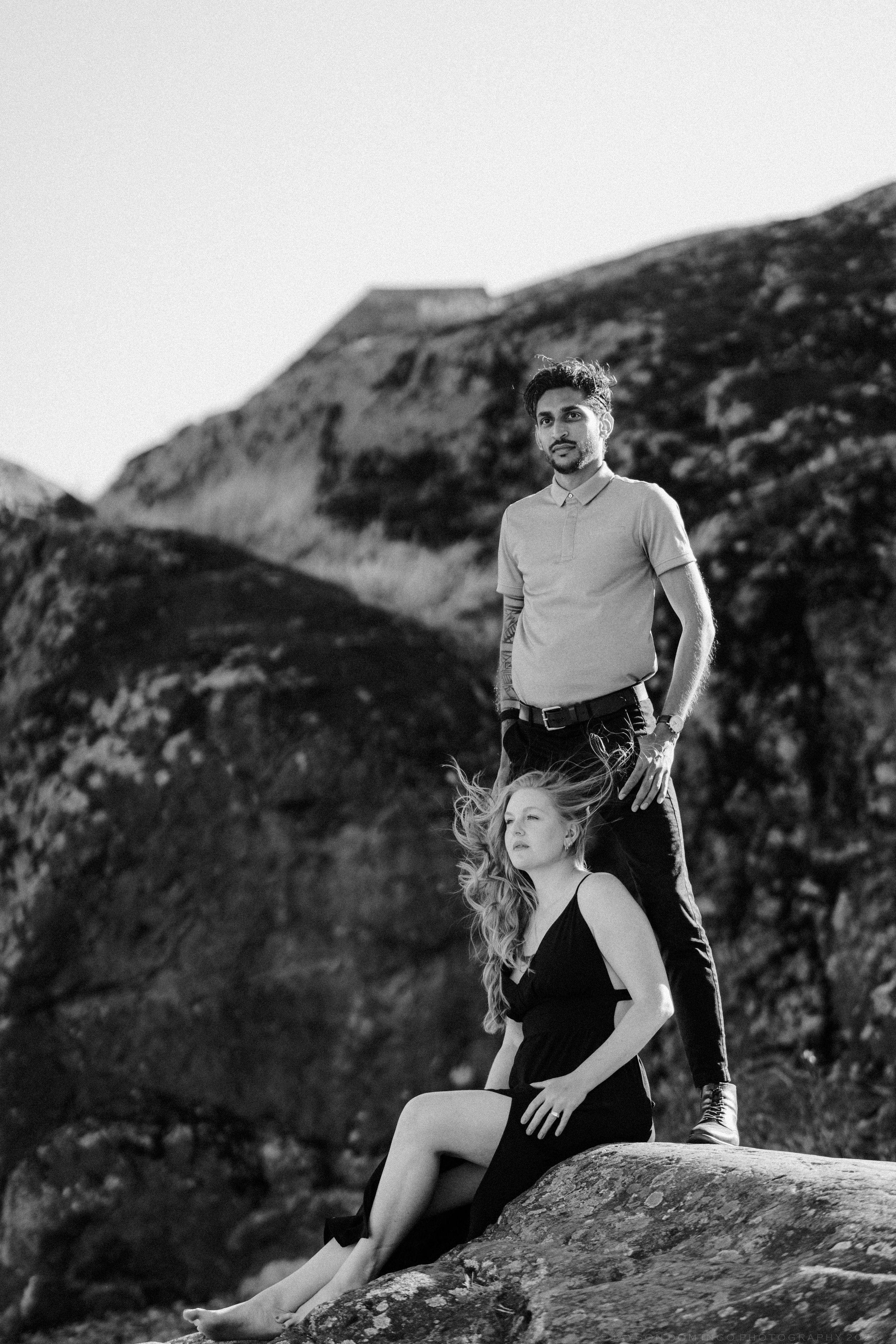 Black and white portrait of a couple on rocks, man standing tall behind seated woman with windswept hair.