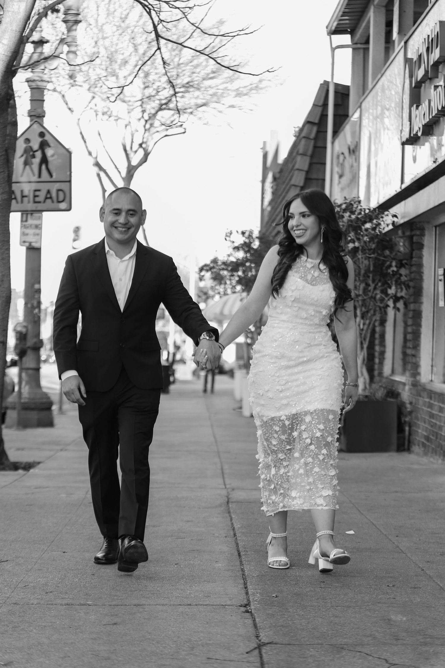 Black and white photo of engaged couple holding hands walking on city sidewalk in formal attire.