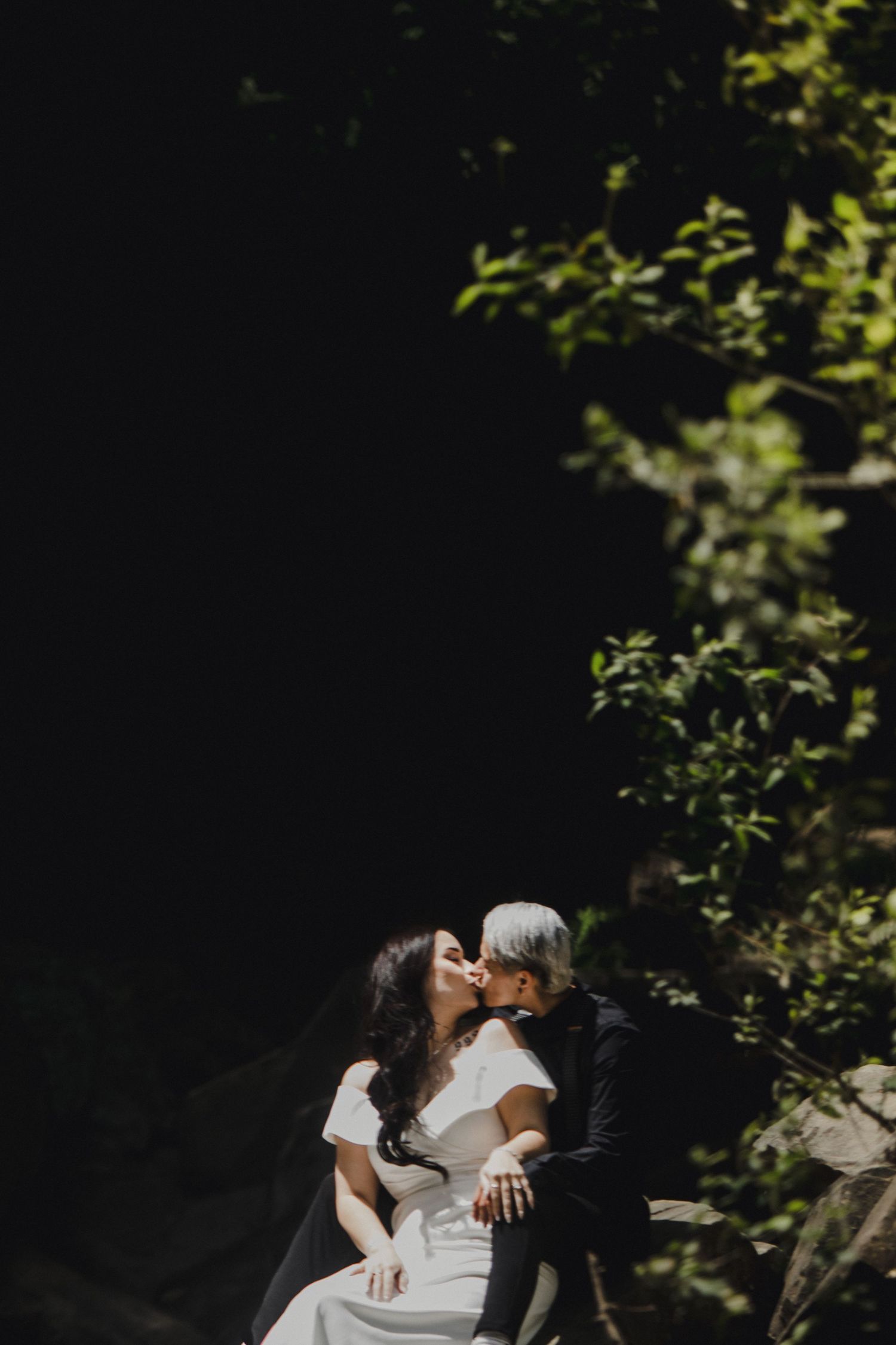 A couple shares a romantic kiss in dappled sunlight, surrounded by lush green foliage, the bride wearing an elegant white gown.