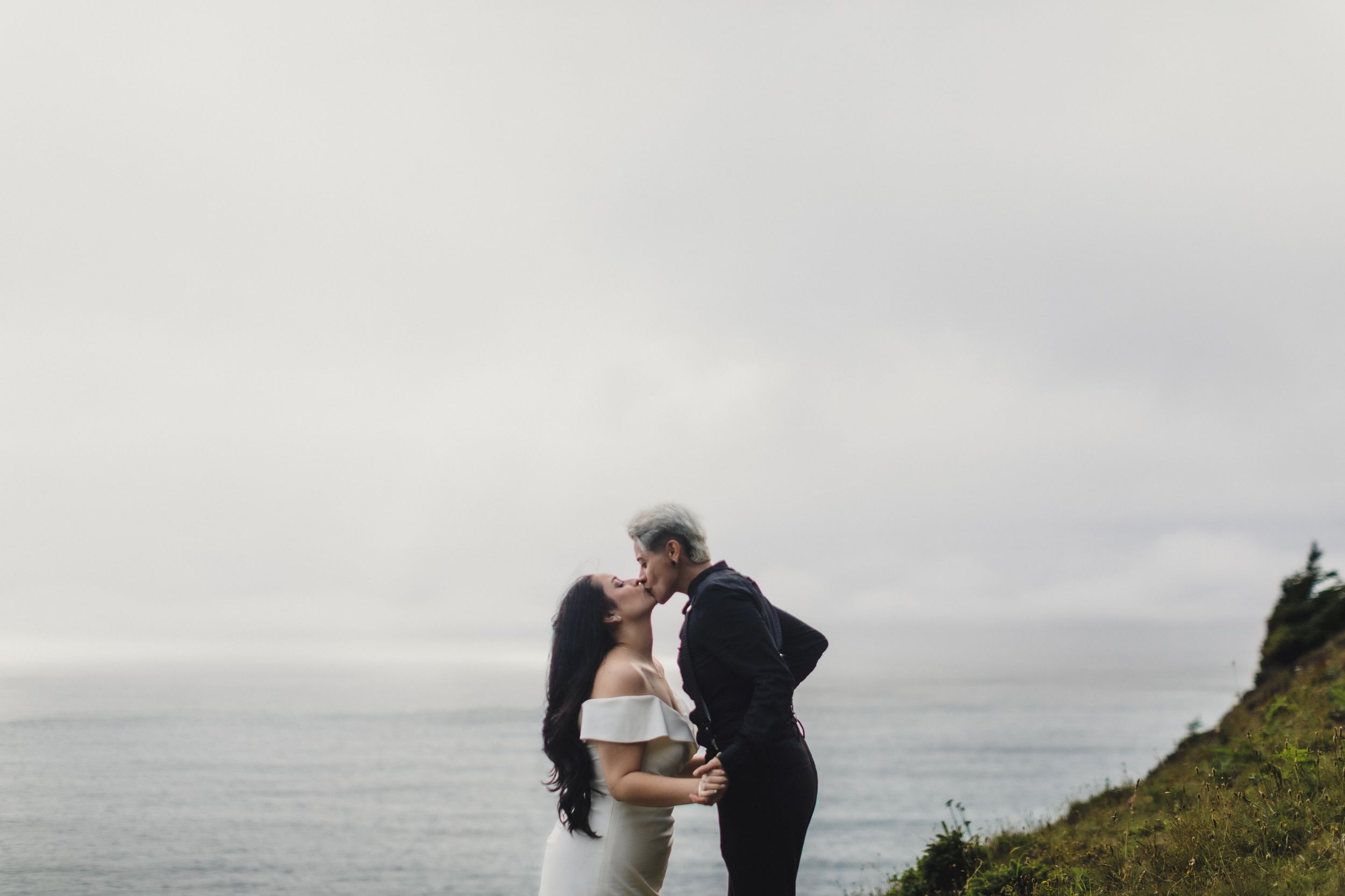 Couple sharing a romantic kiss on a coastal cliff with ocean views on a misty wedding day.