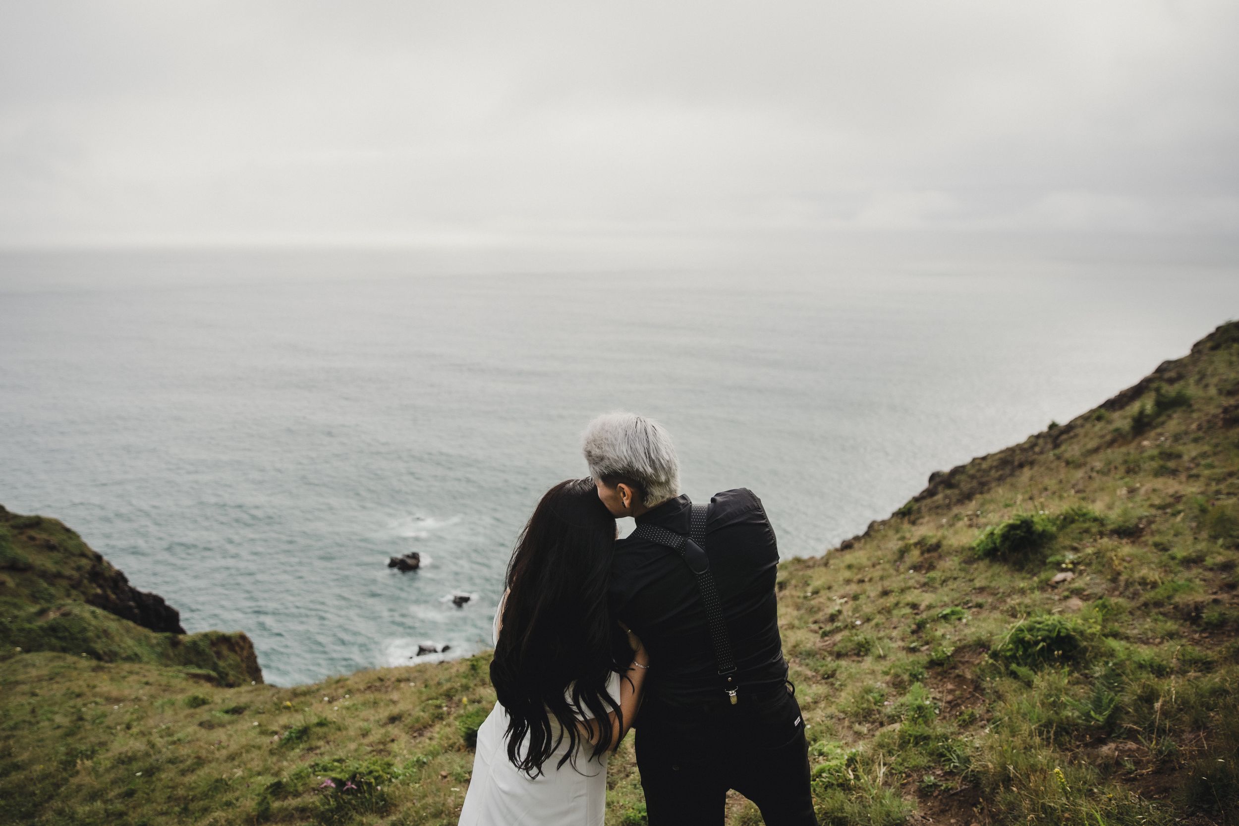 Couple sharing a romantic kiss on a coastal cliff overlooking the misty ocean on their wedding day.