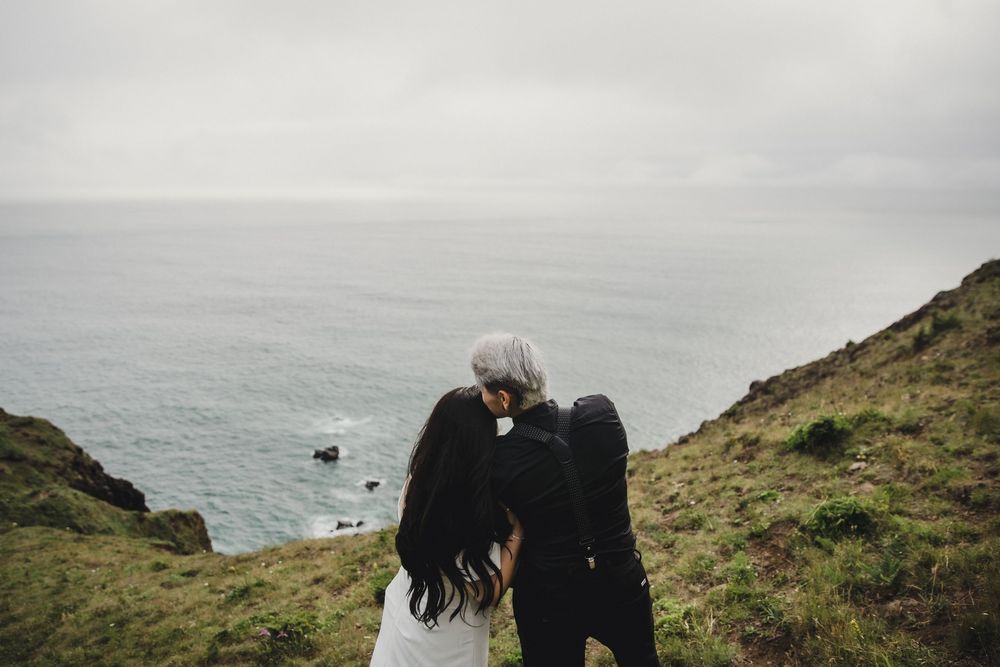 Couple sharing a romantic kiss on a coastal cliff overlooking the misty ocean on their wedding day.
