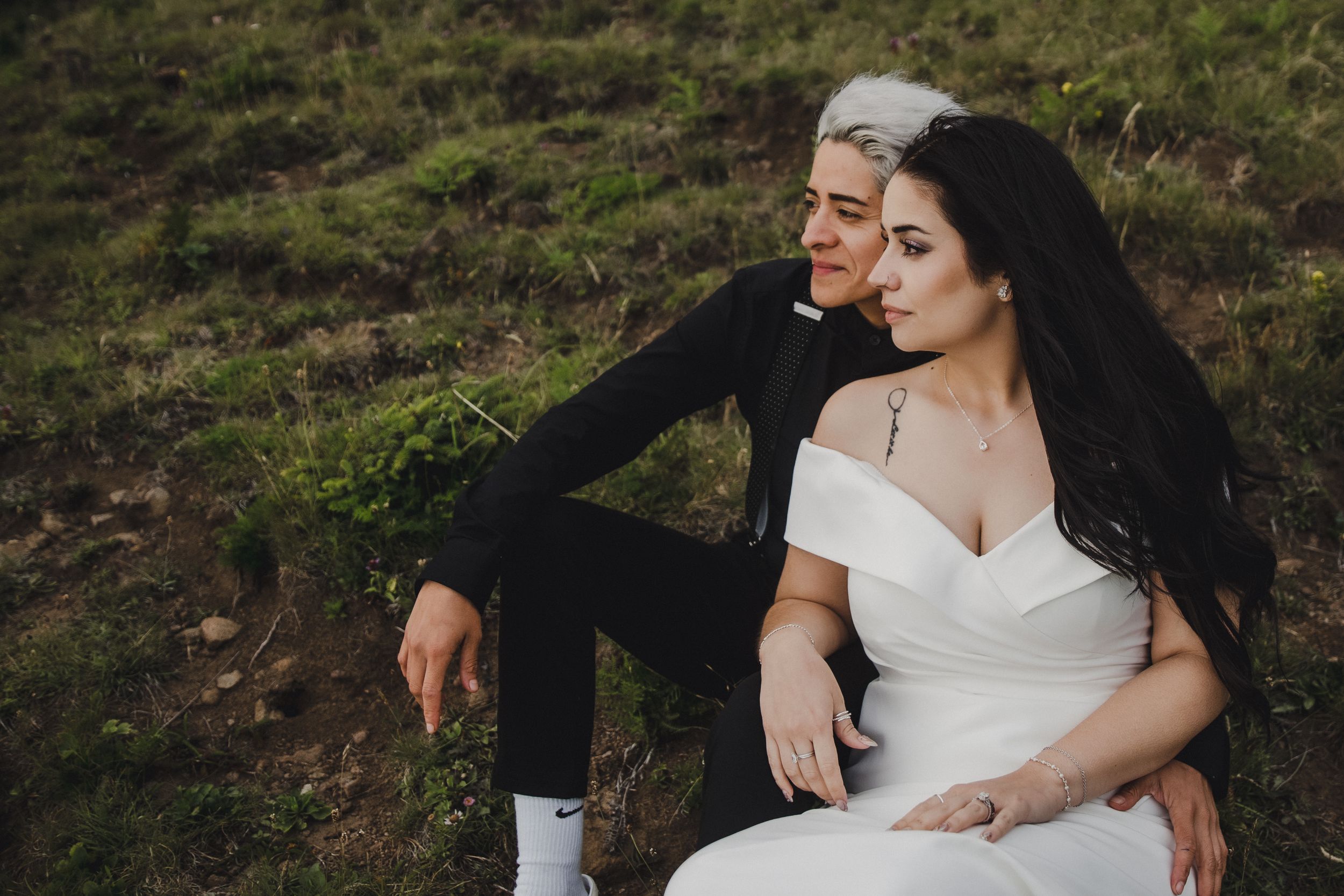 Two brides in wedding attire sit together on a grassy hillside, one in a white gown and one in a black suit.