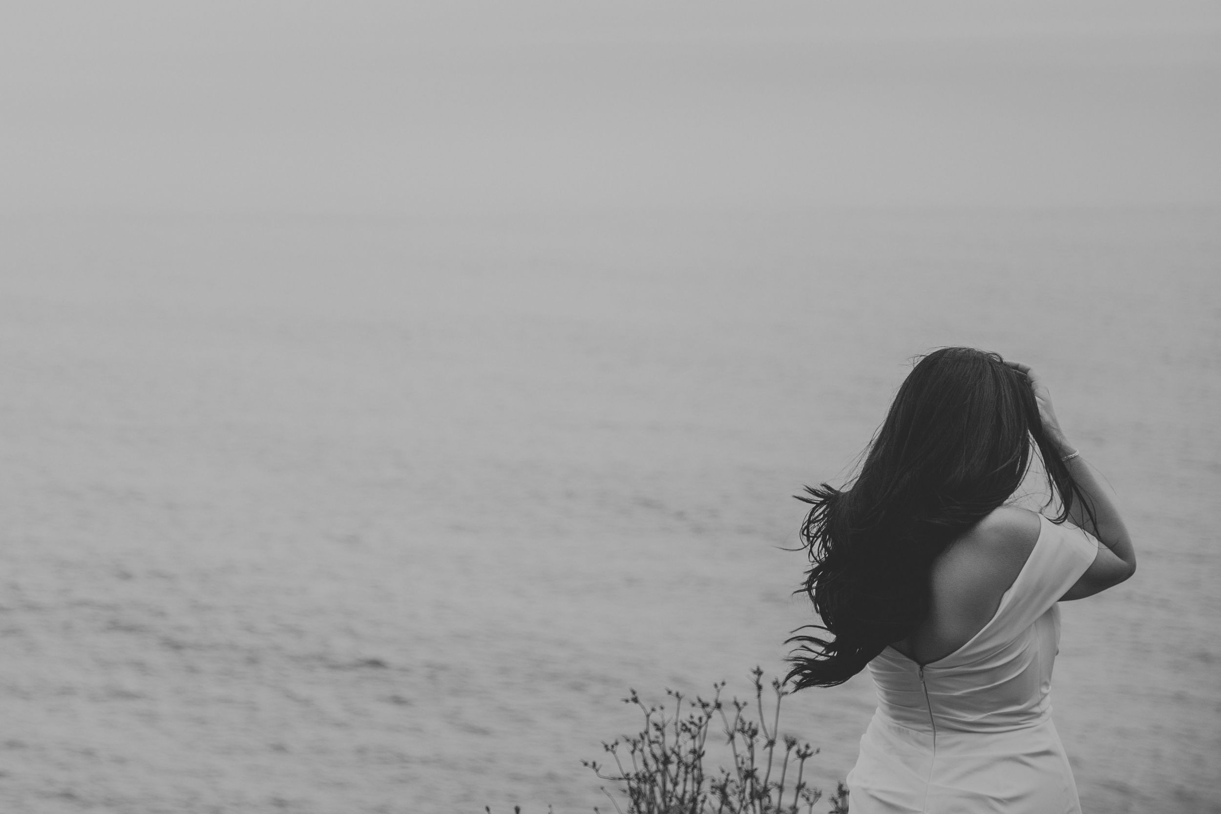 Black and white photo of a woman with long dark hair standing by the ocean, her back turned as wind blows her hair.