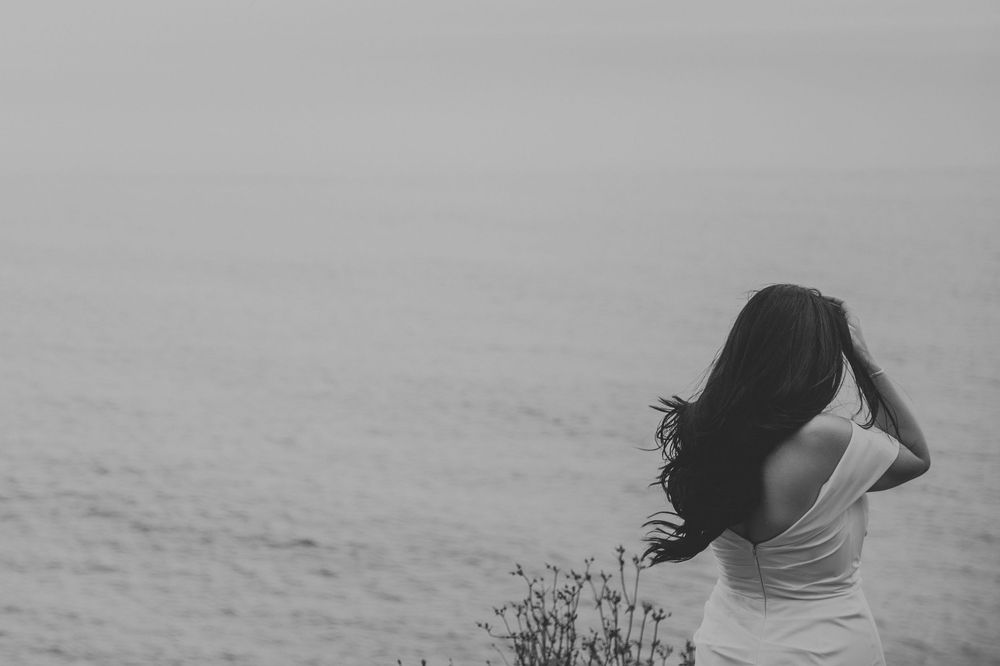 Black and white photo of a woman with long dark hair standing by the ocean, her back turned as wind blows her hair.