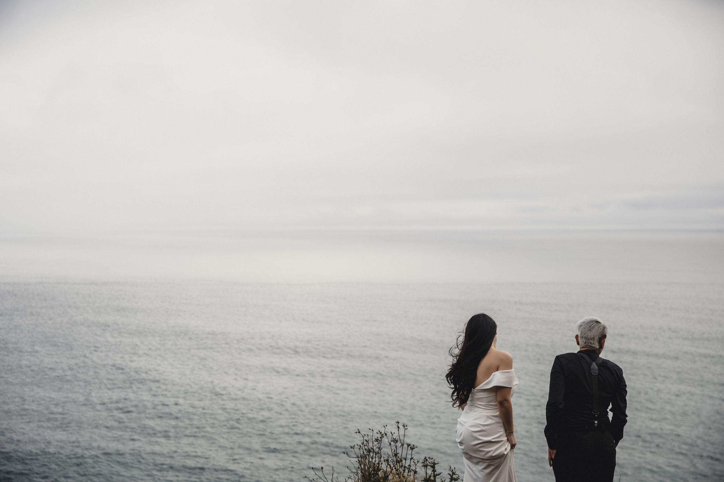 Bride and groom standing on a cliff edge gazing at the calm misty ocean on their wedding day.