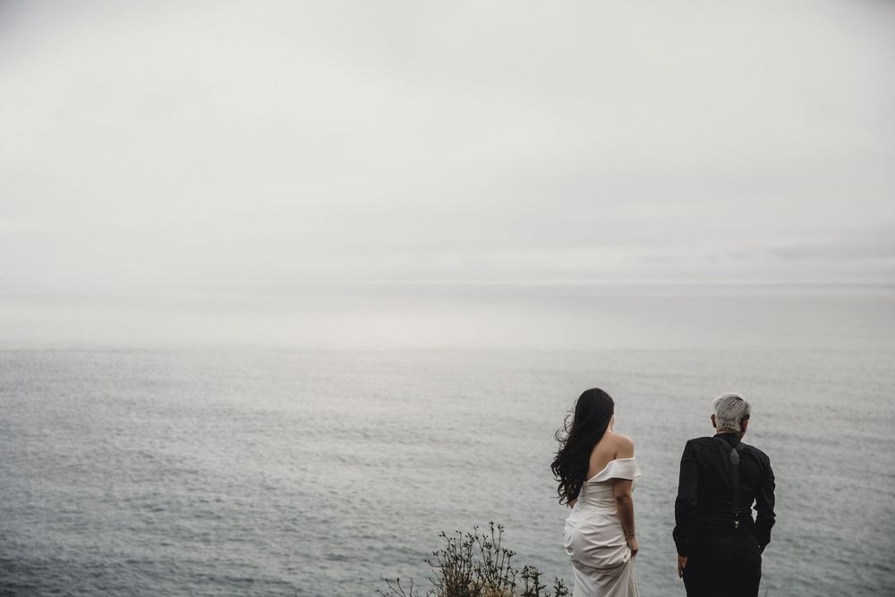 Bride and groom standing on a cliff edge gazing at the calm misty ocean on their wedding day.
