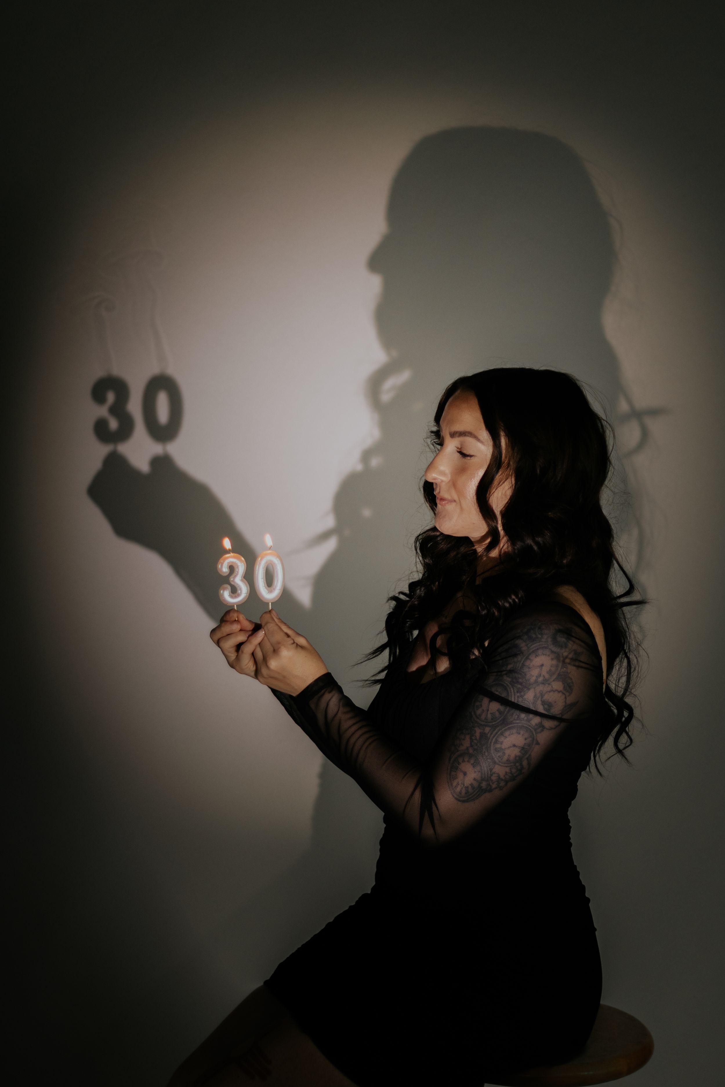 Woman in black lace dress holding sparkler number 30 candles, celebrating 30th birthday with shadow on wall.