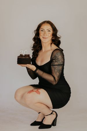 Woman in black dress crouching while holding a chocolate birthday cake with a 30 candle, smiling at camera.