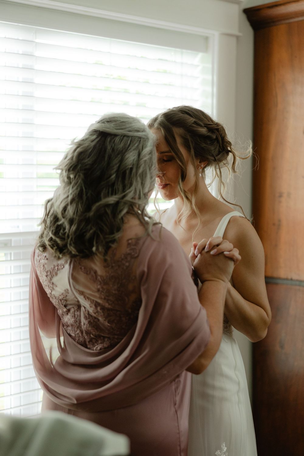 A bride in a white gown holds hands with a woman in a pink dress by a sunlit window.