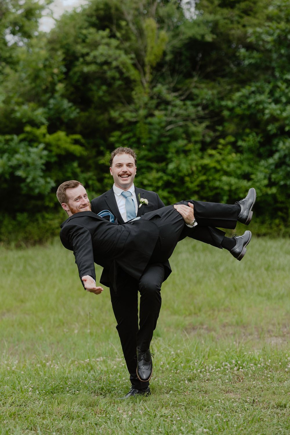 Groomsman playfully carries groom in black suit across a grassy field at outdoor wedding.