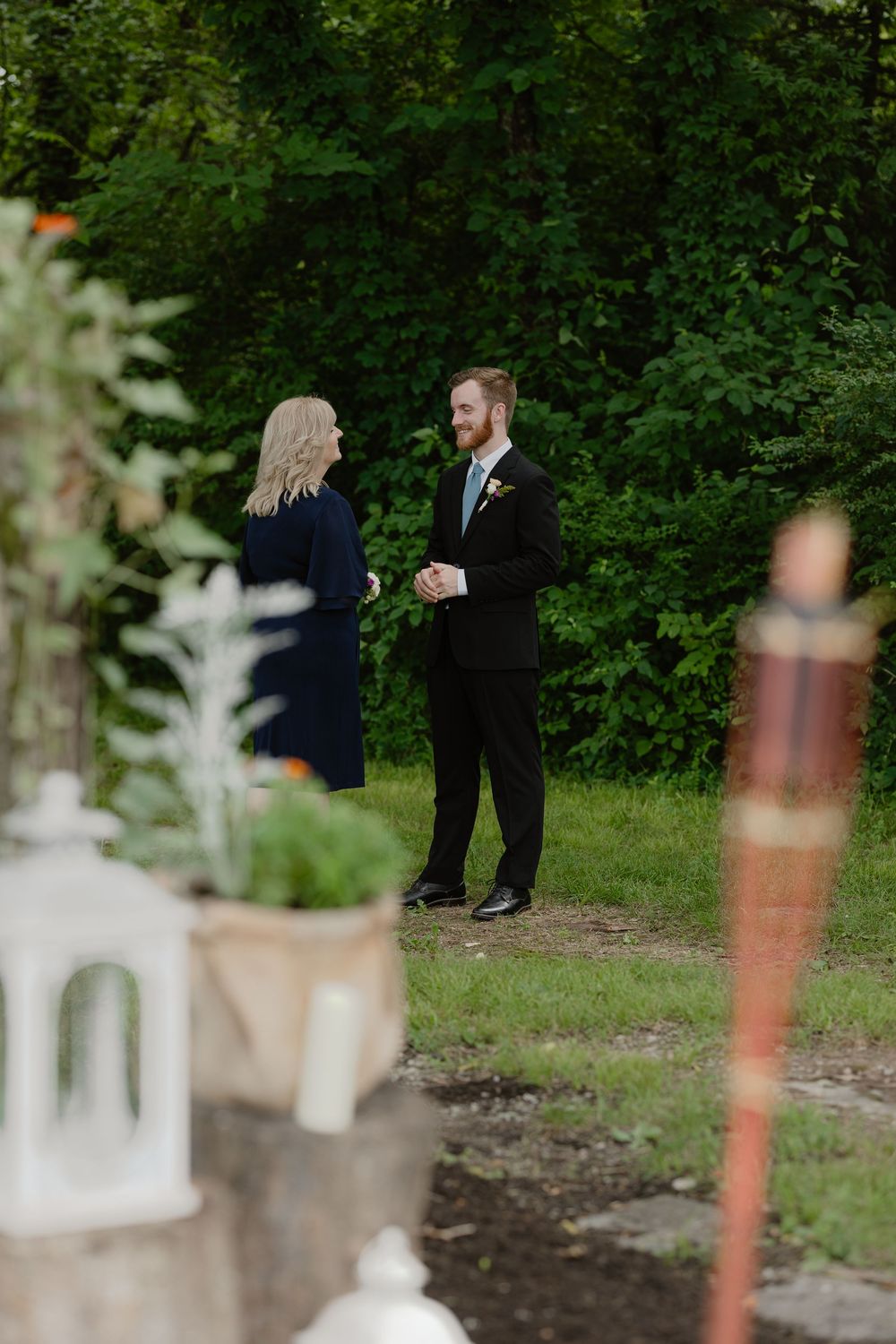 A groom in a black suit with boutonniere speaks with a woman in a navy dress near floral arrangements.