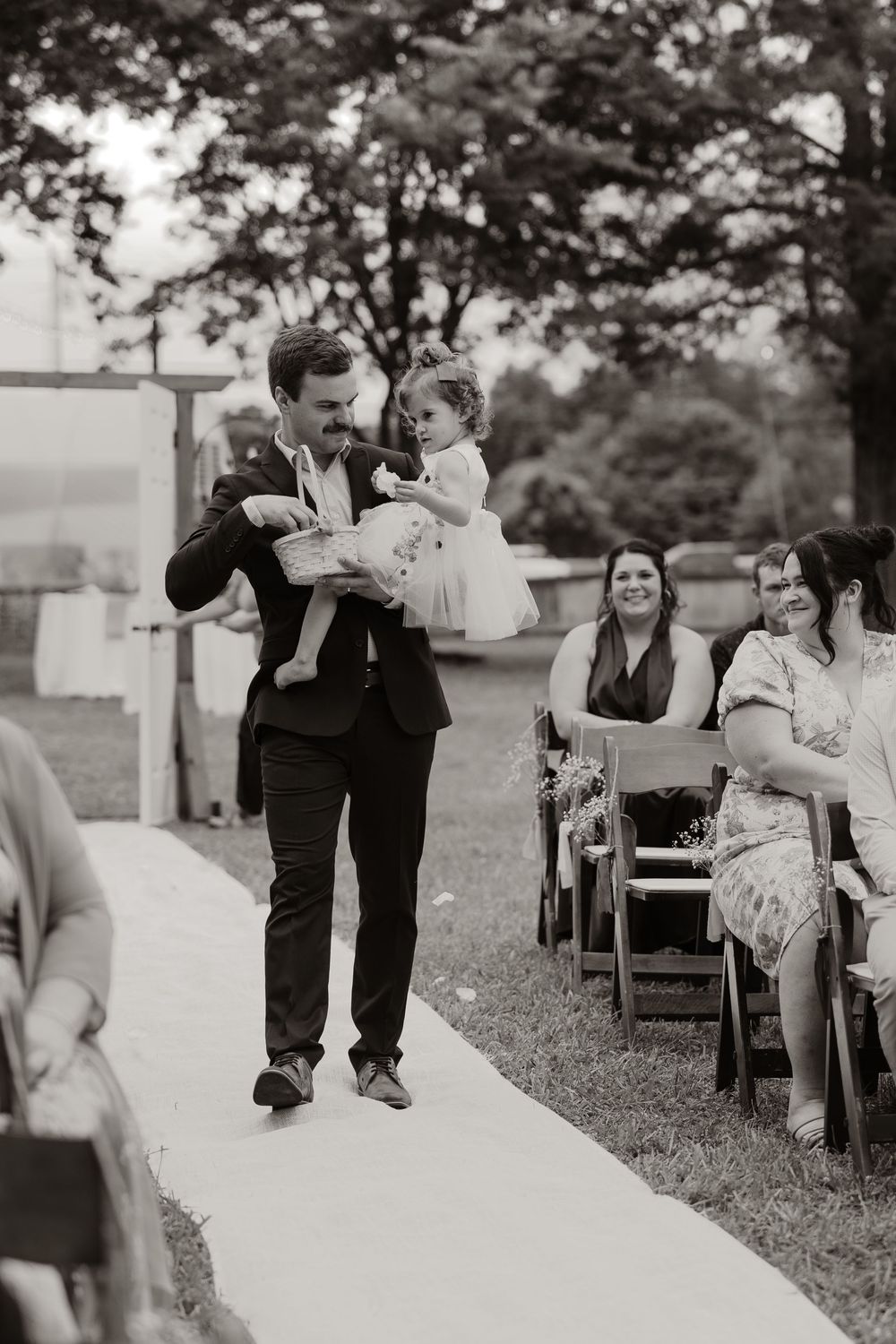 Black and white photo of groomsman carrying young flower girl down outdoor wedding ceremony aisle.