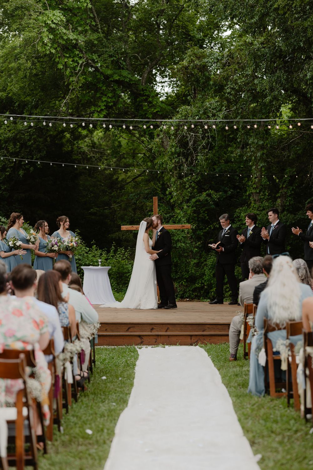 Bride and groom share first kiss on wooden stage at outdoor garden wedding with string lights.
