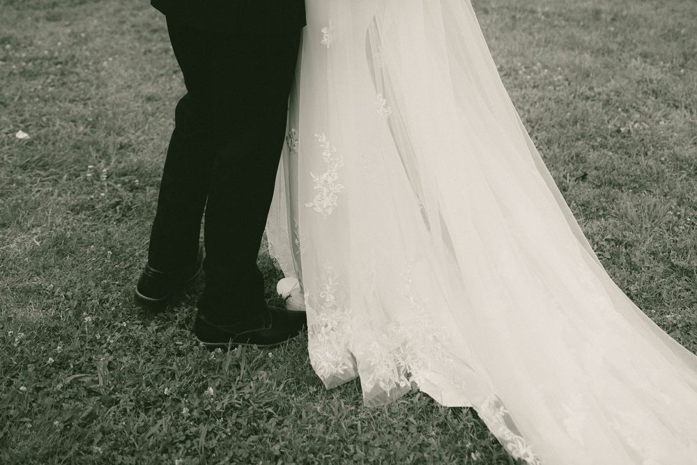 Black and white close-up of bride in lace wedding gown standing beside groom on grass.