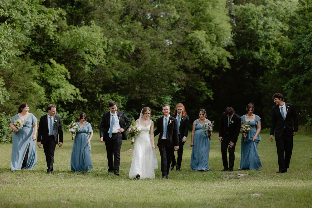Wedding party walks together across green lawn, bridesmaids in dusty blue gowns and groomsmen in black.