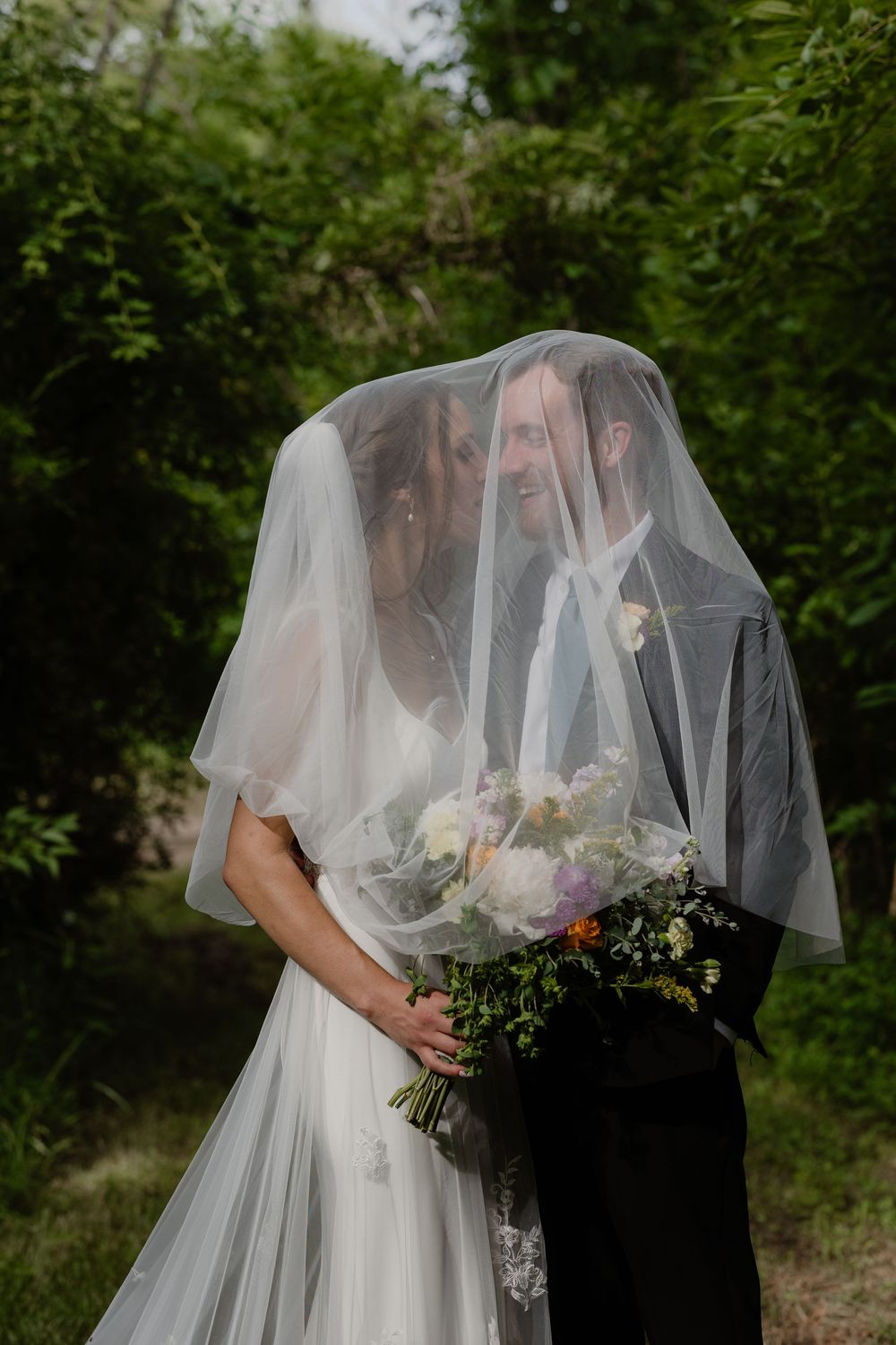 Bride and groom share a hidden kiss beneath the bride's flowing veil holding a floral bouquet.