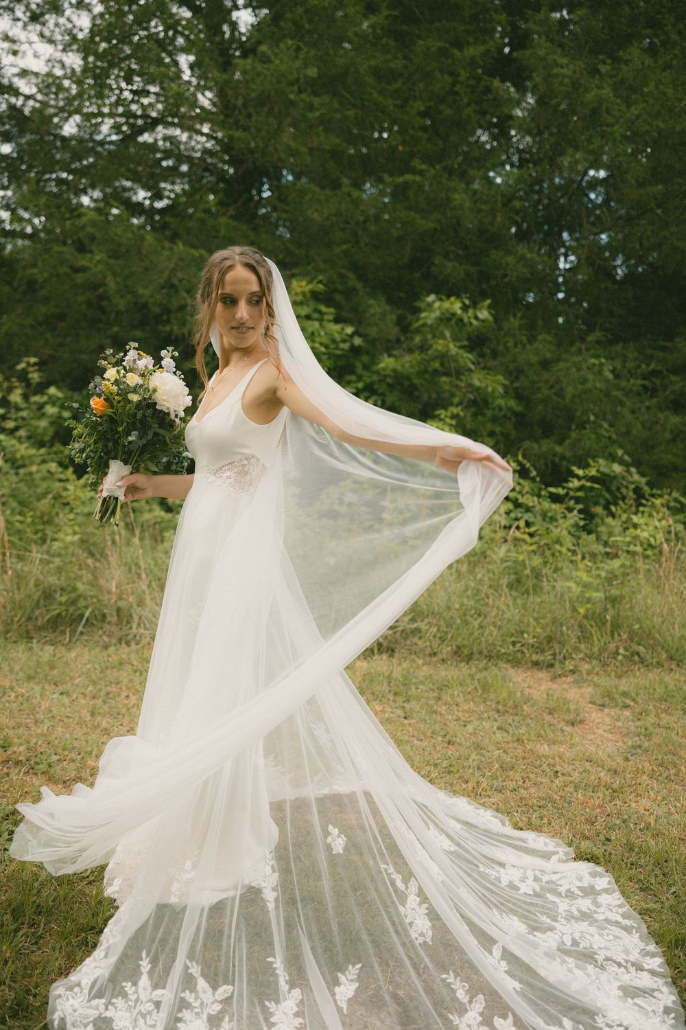 Elegant bride in lace wedding gown twirling her veil while holding a bouquet in a natural green landscape.