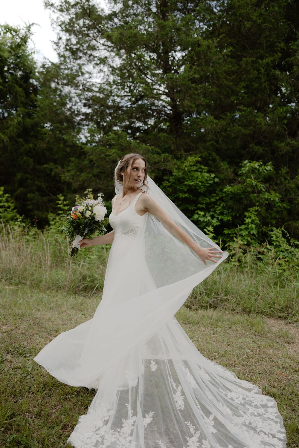 Beautiful bride in lace wedding dress lifting her cathedral veil while posing with bouquet in a green field.