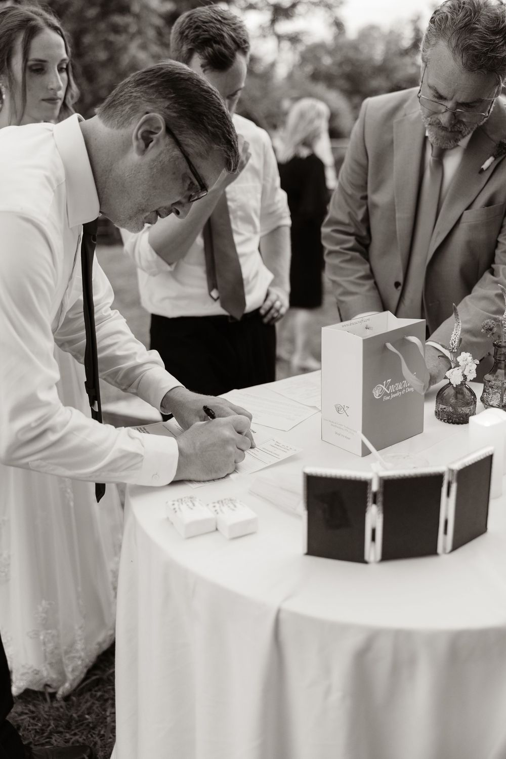 Groom signs wedding documents at a white tablecloth table during an outdoor wedding ceremony.