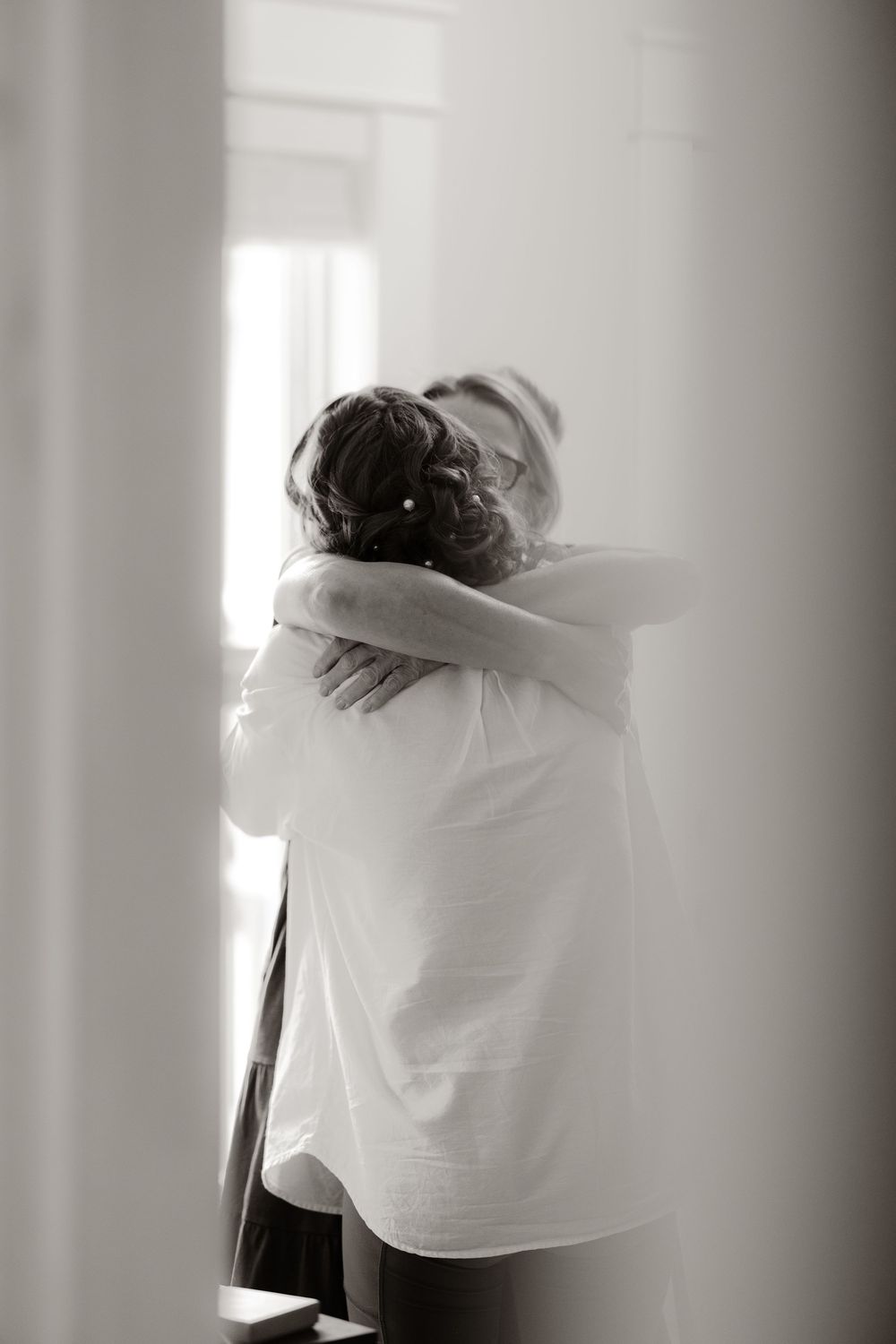 Emotional black and white photo of bride in white shirt sharing a tender hug near a bright window.