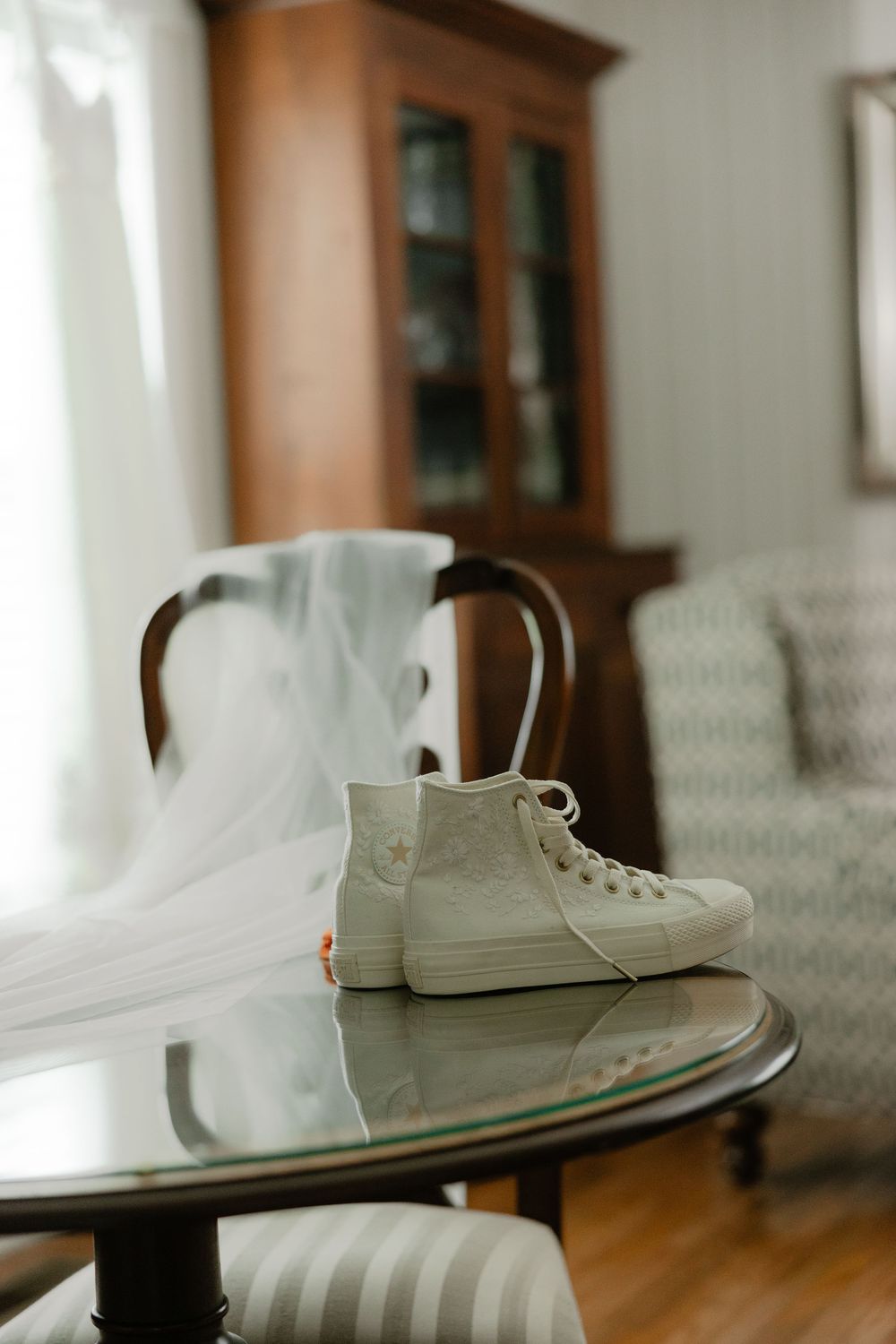 White Converse high-top sneakers on a glass table with a bridal veil draped over a chair.