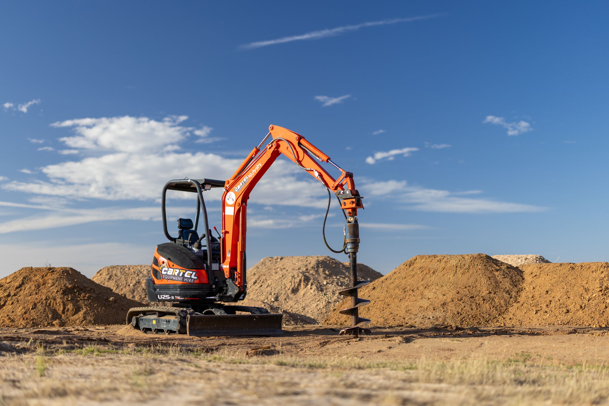 Orange Kubota mini excavator with auger attachment on a dry dirt field with blue sky backdrop.