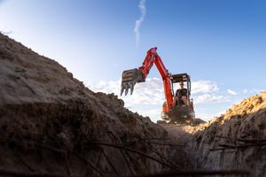 Orange mini excavator digging a deep trench in sandy soil against a bright blue sky.