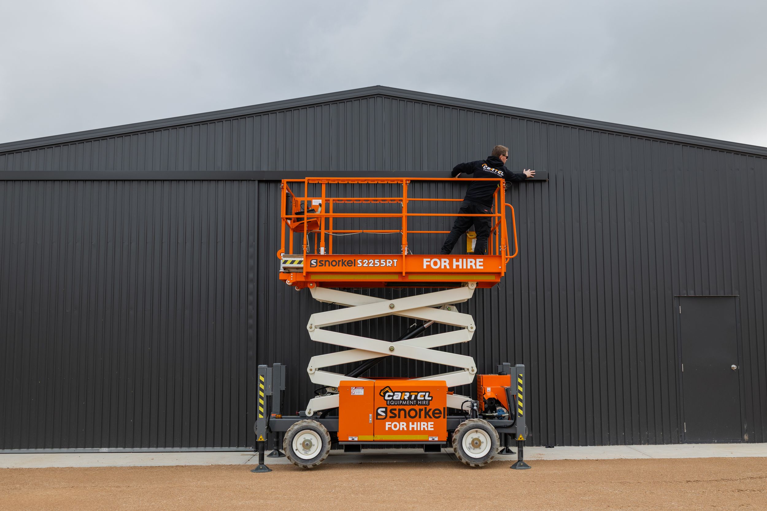 Orange scissor lift elevated against a large dark grey industrial shed wall with hire signage.