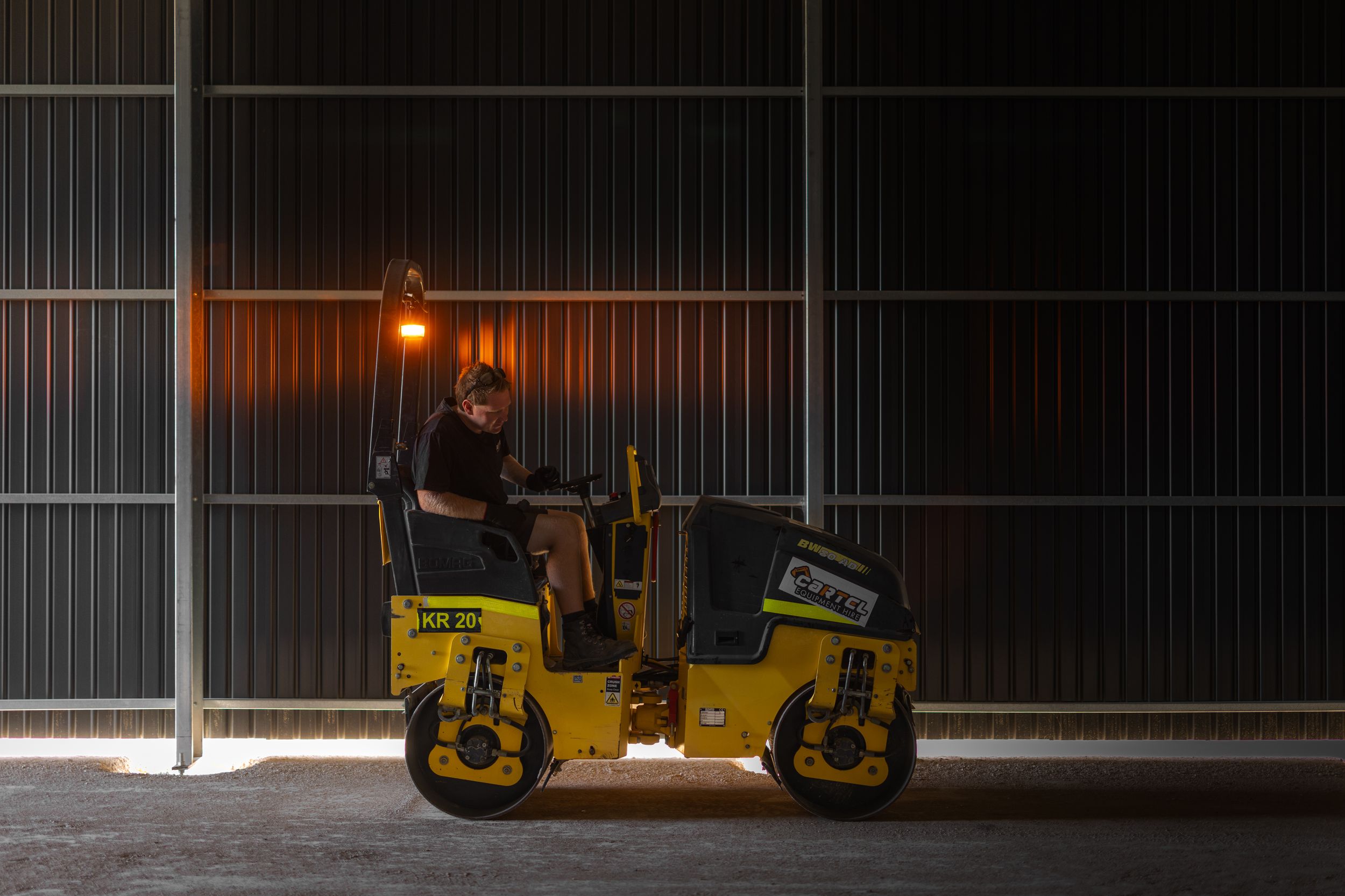 Worker operating a yellow road roller compactor at night on a construction site.