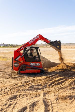 Red Manitou tracked skid steer loader dumping sand on a construction site under blue sky.