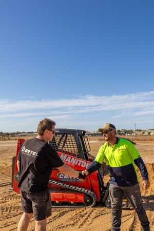 Two construction workers shaking hands in front of a red Manitou skid steer loader.