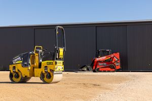 Yellow road roller and red Manitou skid steer parked outside a dark industrial building.