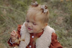 Adorable toddler girl with bow in hair wearing rust dress and sherpa vest, smiling outdoors in fall.